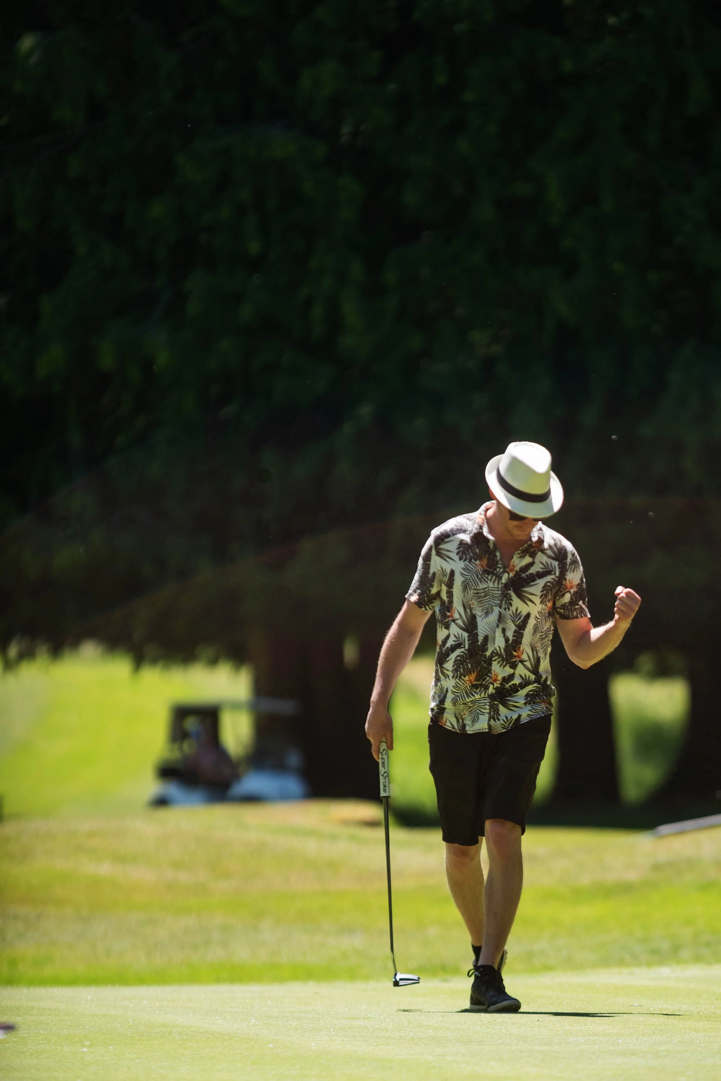 A man in a tropical shirt, shorts, and a white hat is walking on a golf course holding a golf club, with a golf ball and tee in front of him, and trees in the background.