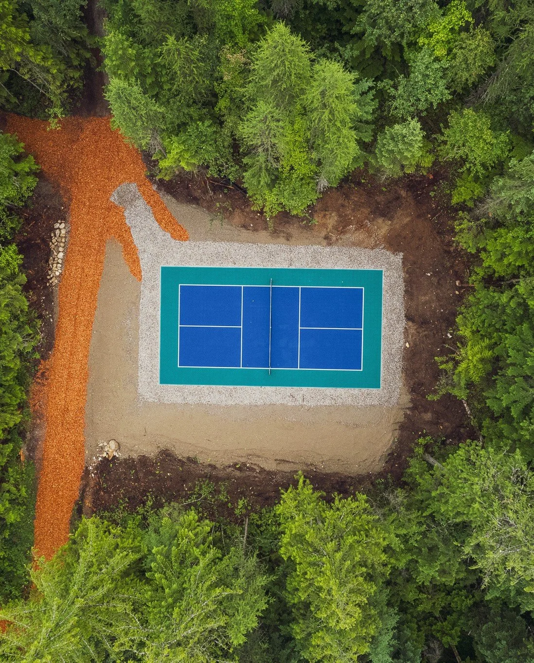 An aerial view of a tennis court surrounded by trees, with a gravel pathway on one side.
