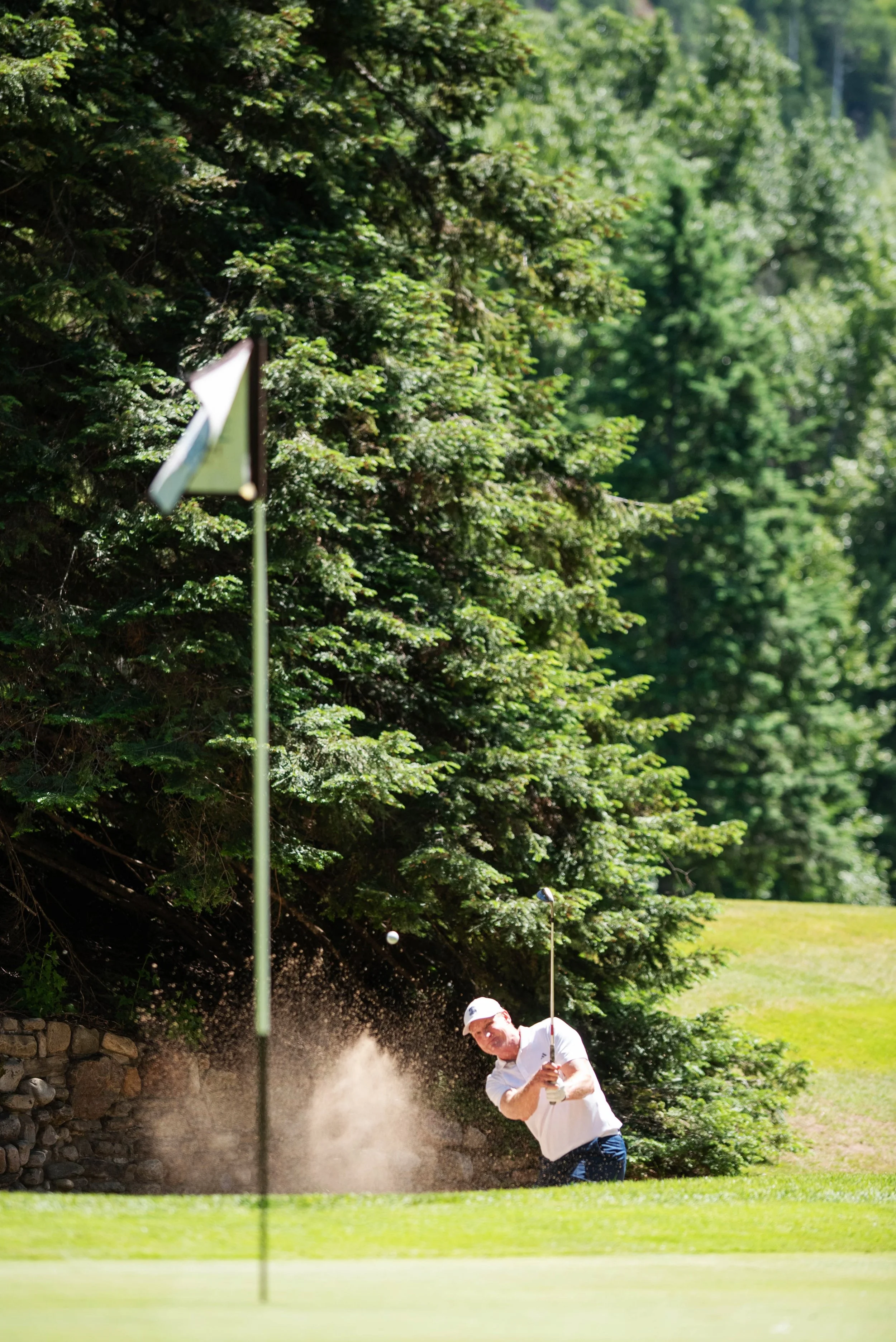 A man wearing a white shirt and a white cap is hitting a golf ball out of a sand bunker on a golf course, with a penalty flag and trees in the background.