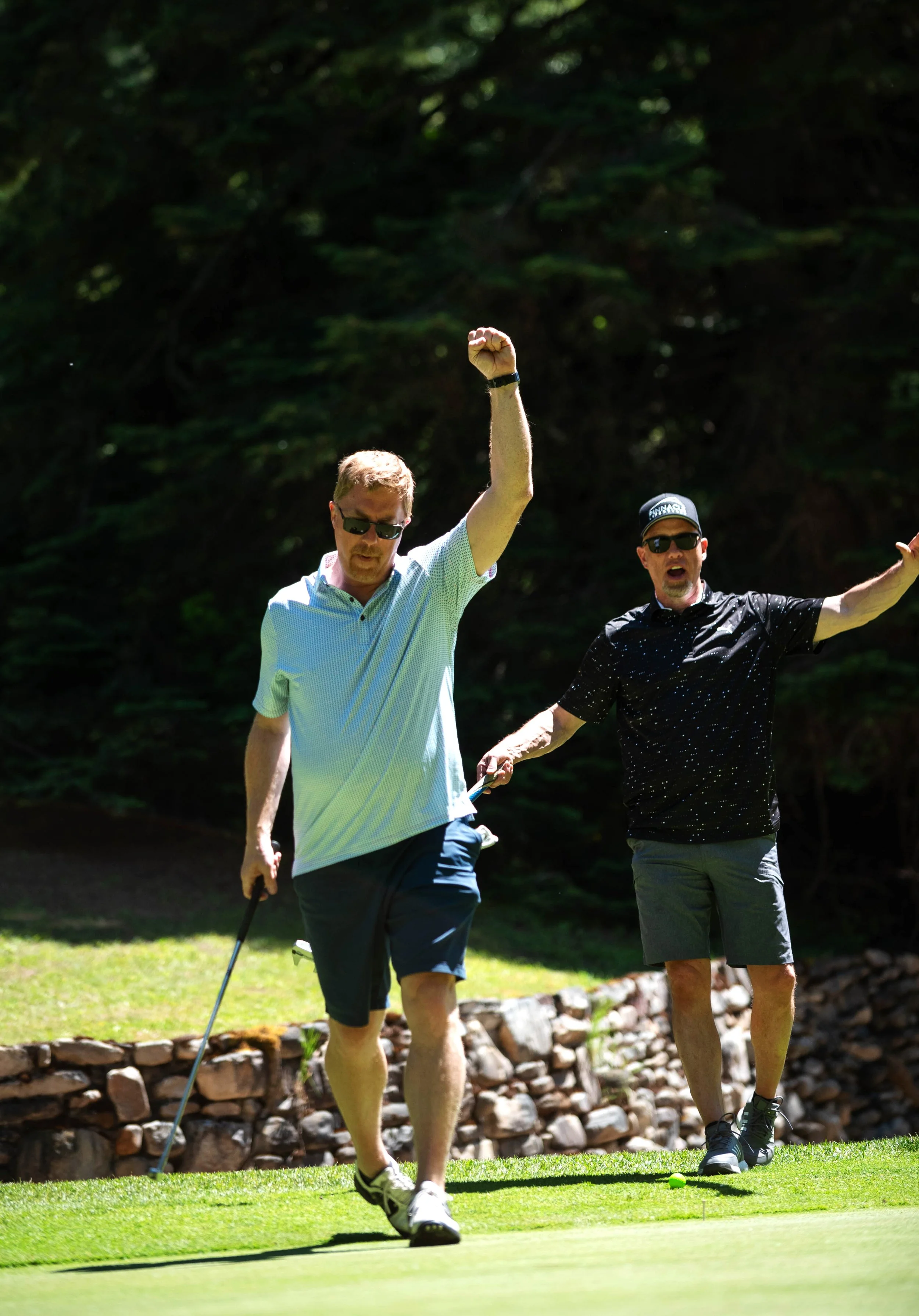 Two men playing golf on a sunny day, one walking with a golf club and the other preparing to hit a golf ball, surrounded by trees and a stone border.