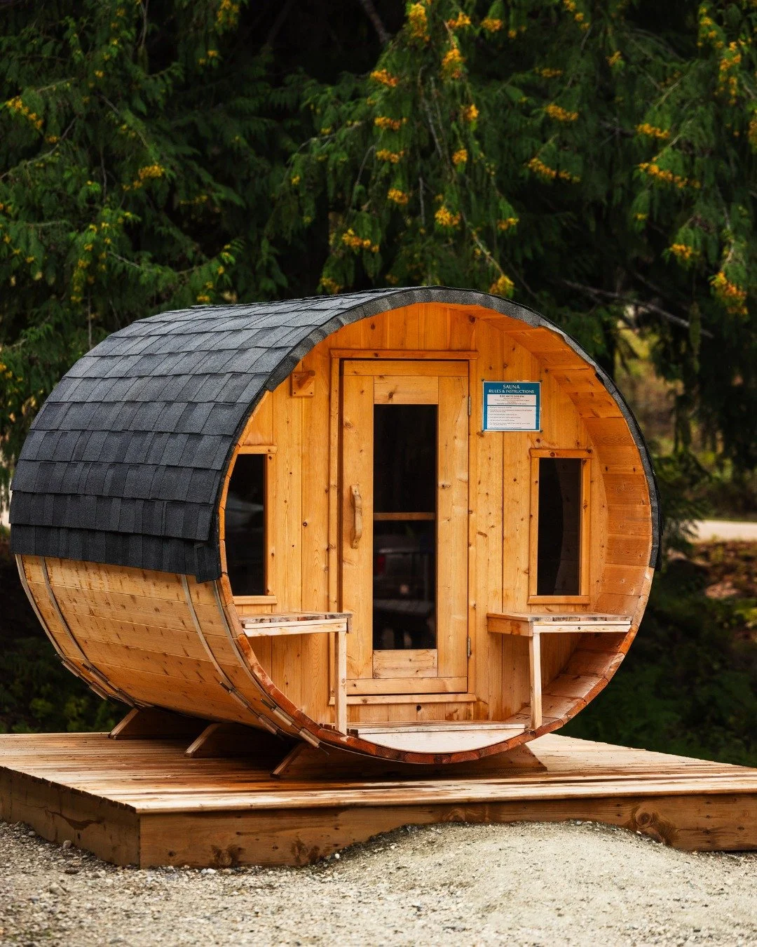 A small, cylindrical wooden sauna with a curved roof, situated on a wooden deck outdoors with trees in the background.