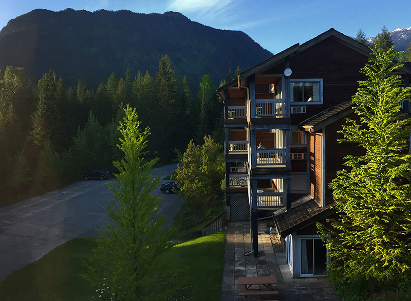 A multi-story wooden lodge with balconies, surrounded by green trees and mountains in the background, during daylight.