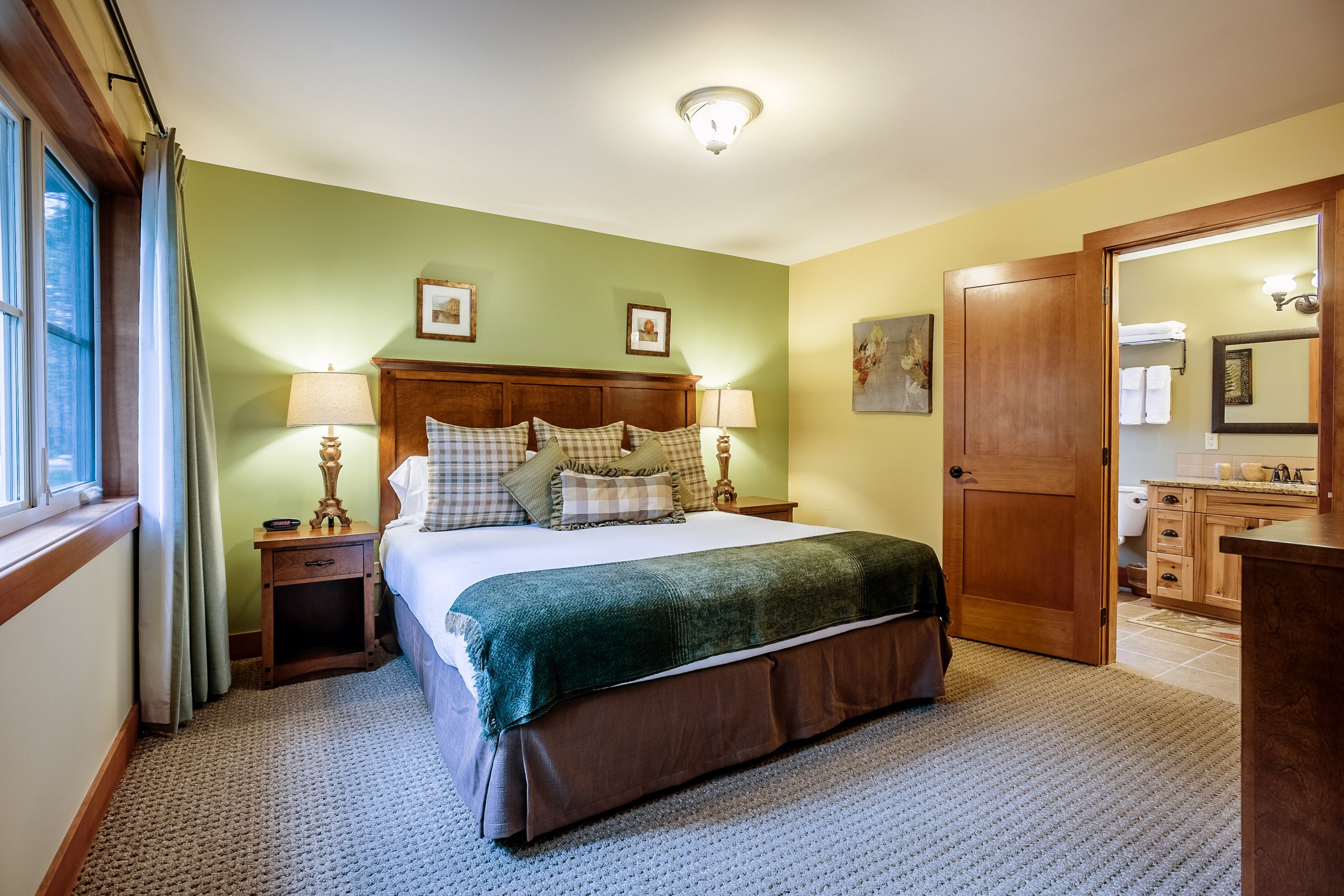 A cozy bedroom with a wooden headboard, checkered pillows, lamps, framed artwork, and a view into a bathroom with towels and mirror.