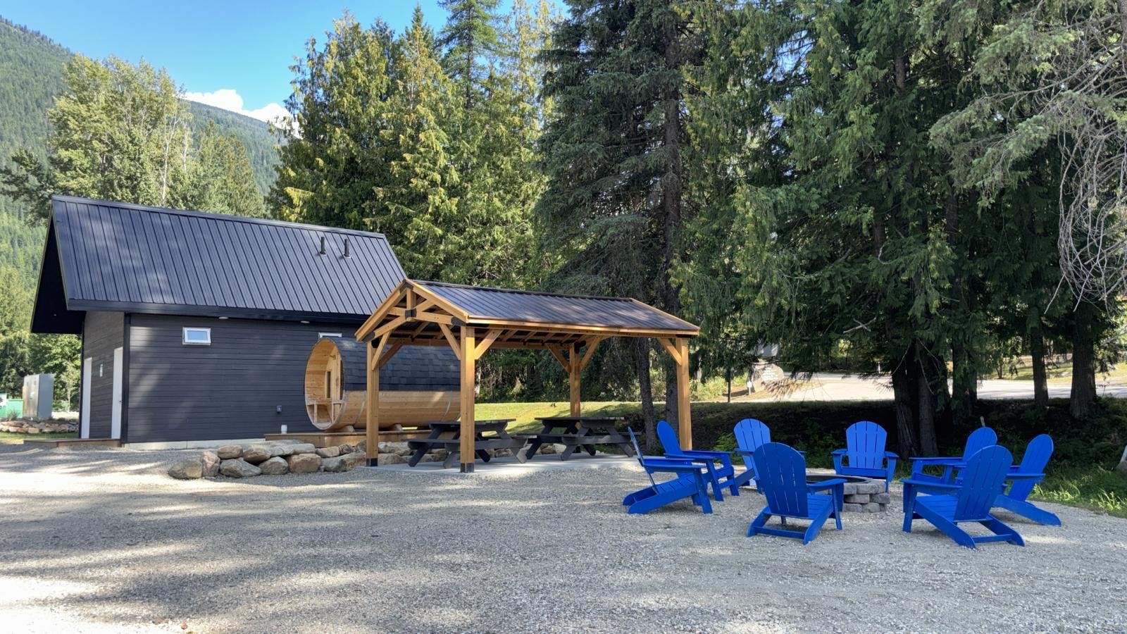 A small dark gray building with a rounded wooden porch and a circular window, surrounded by green trees, with a gravel area in front featuring six blue Adirondack chairs arranged in a semi-circle and picnic tables beneath a wooden shelter.