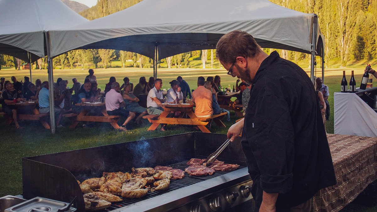 A man grilling steaks and chicken at an outdoor barbecue during a gathering, with people sitting under a large canopy in the background, on a grassy field surrounded by trees.