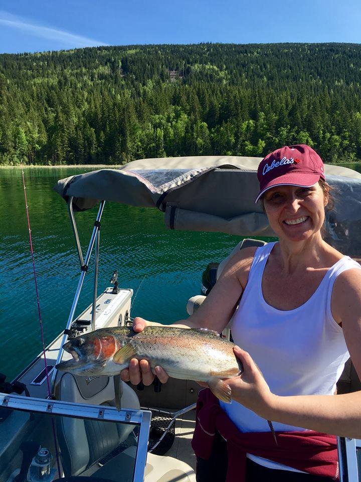A woman on a boat holding a fish she caught, with a backdrop of a green forested hill and a blue sky.