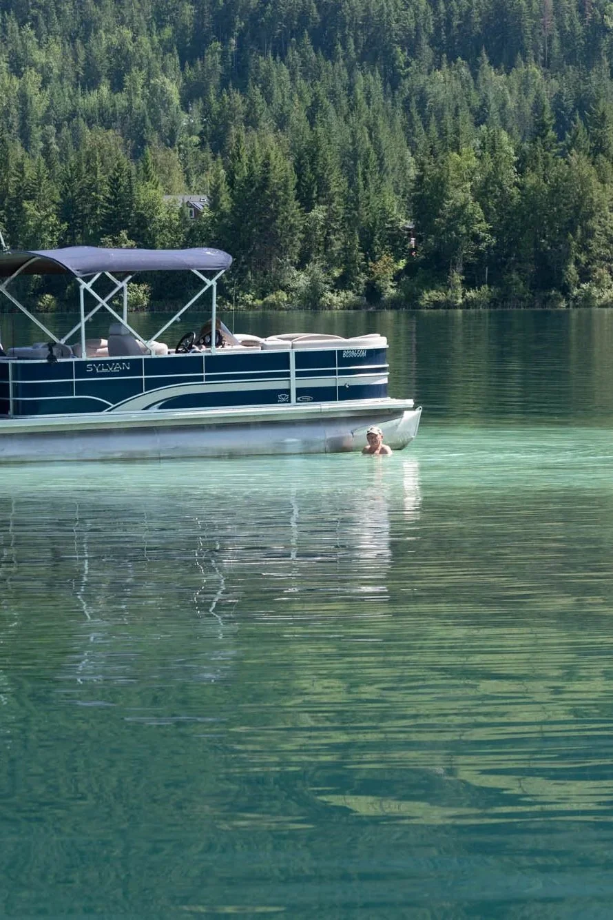 Person swimming near a blue pontoon boat on a calm lake with a background of green forested hills.