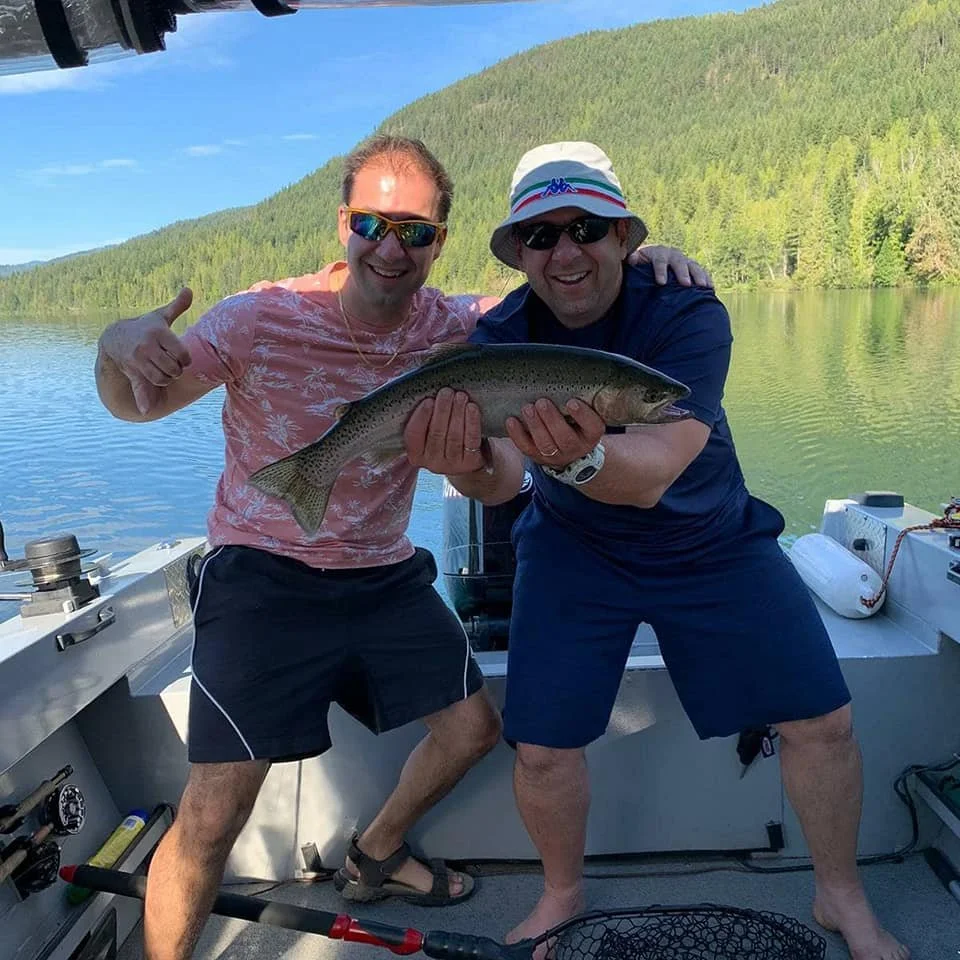 Two men on a boat holding a large fish, with a scenic lake and green hills in the background. One man is giving a thumbs up, both are smiling and wearing sunglasses.