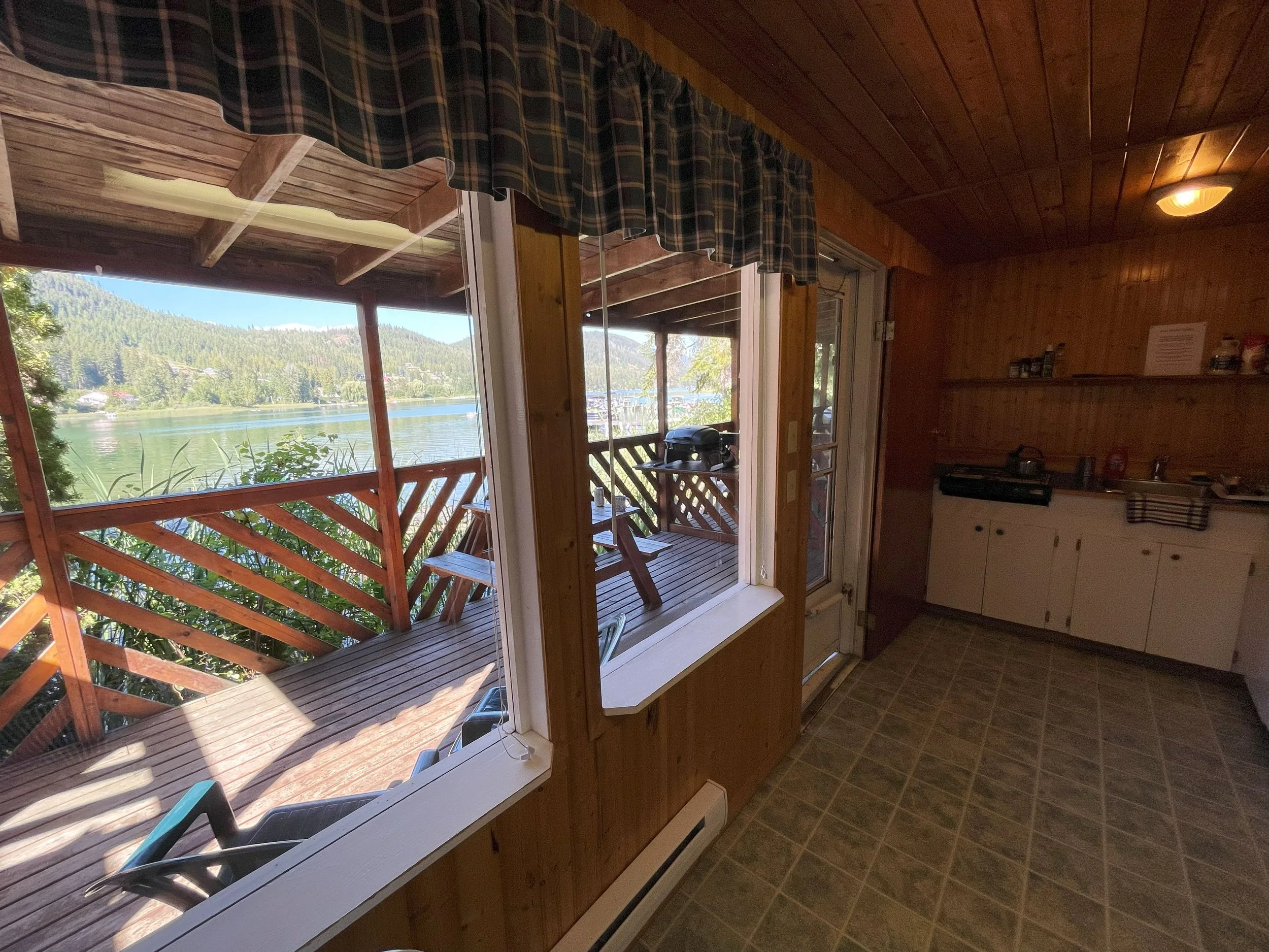 View from inside a cozy wooden cabin looking out onto a balcony with a lake and lush mountains in the background.