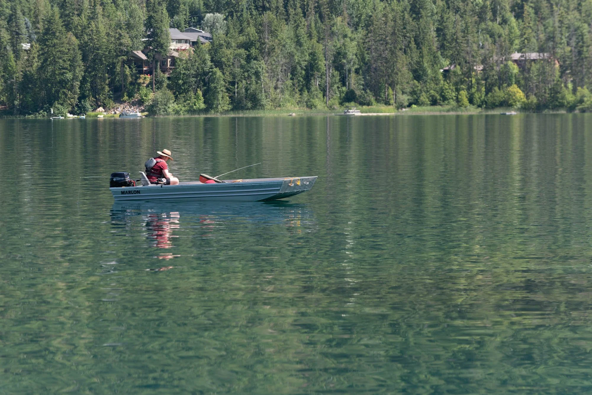 A person fishing on a small boat on a calm lake with a forested shoreline and houses in the background.