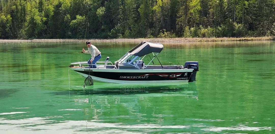 A man standing on a small boat in a green lake, fishing with a fishing rod.