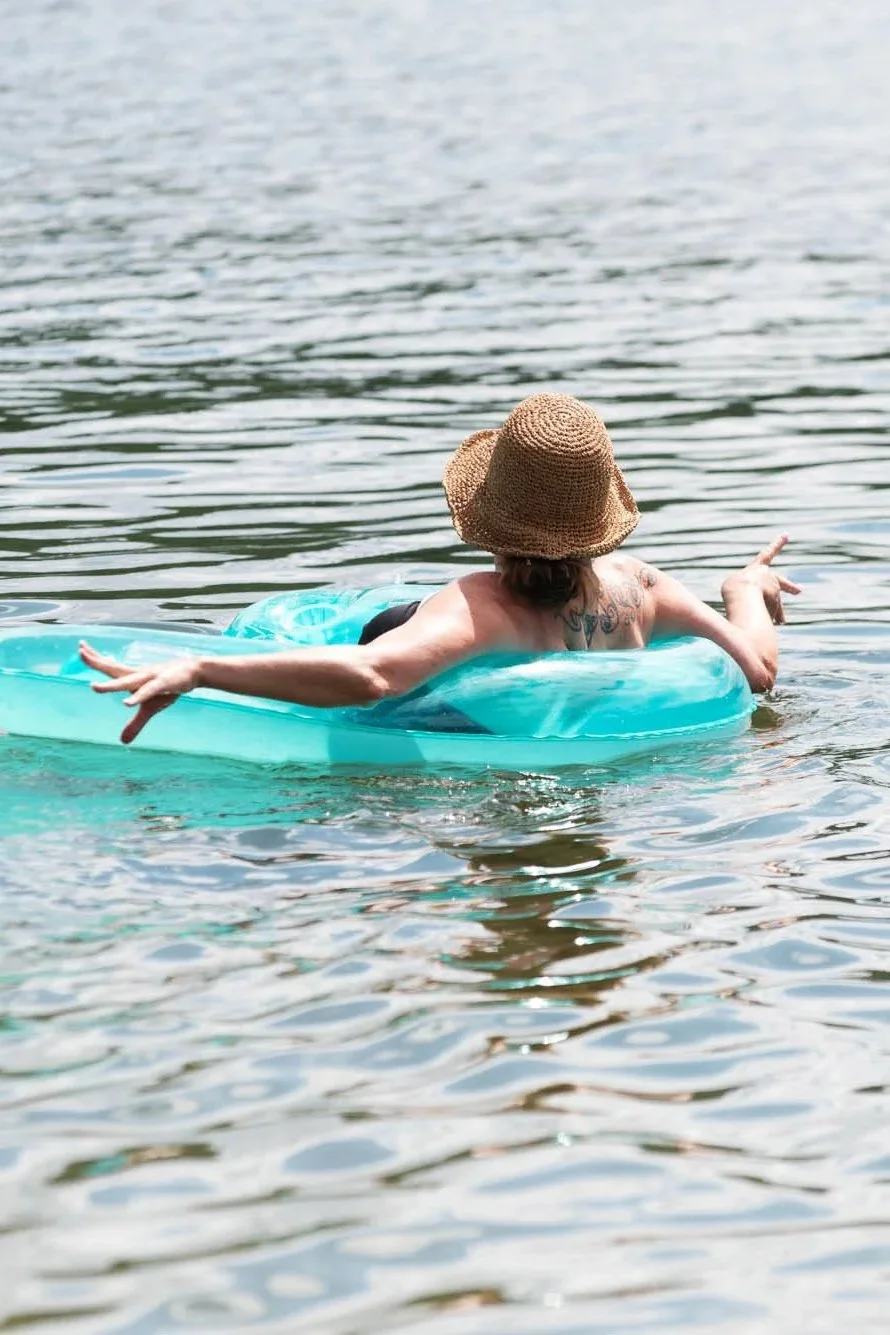 Person floating on a blue pool float in a lake, wearing a wide-brimmed straw hat and black bathing suit, with arms outstretched.