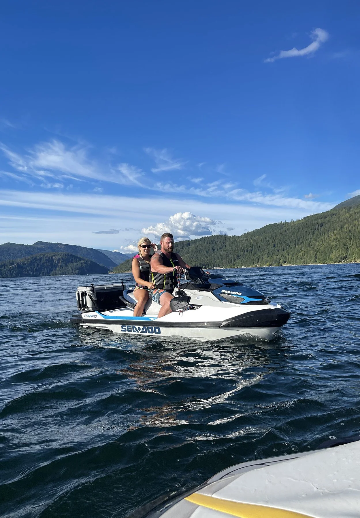 Two people riding a white Sea-Doo jet ski on a lake with mountains and green forest in the background under a blue sky.