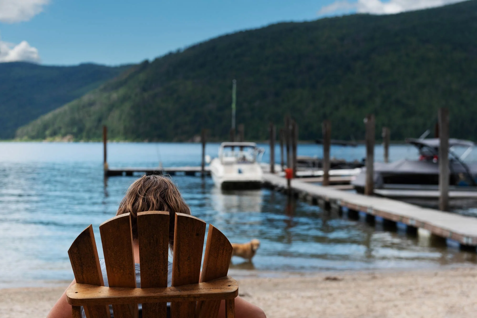 Person relaxing on a wooden chair on a lakeside beach, overlooking a dock with boats and a green hill in the background.