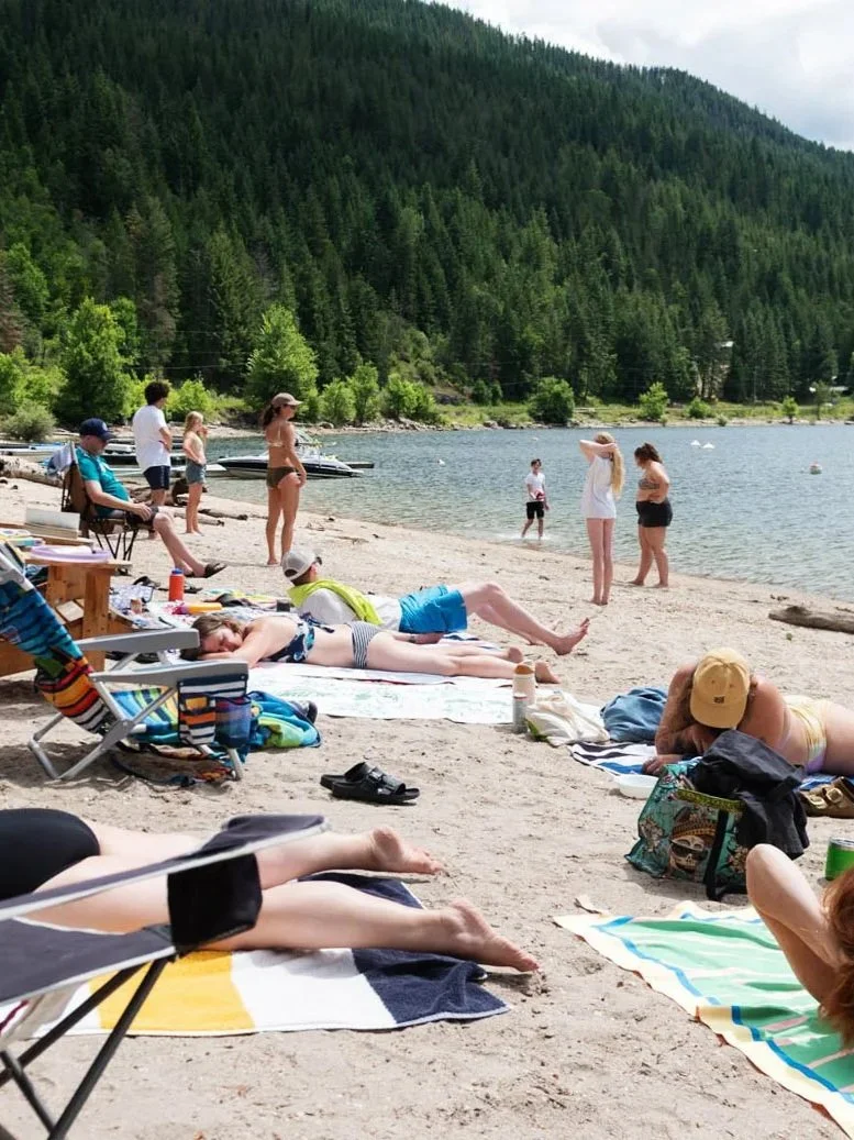 People relaxing and enjoying a day at the beach near a lake with a backdrop of forested mountains.