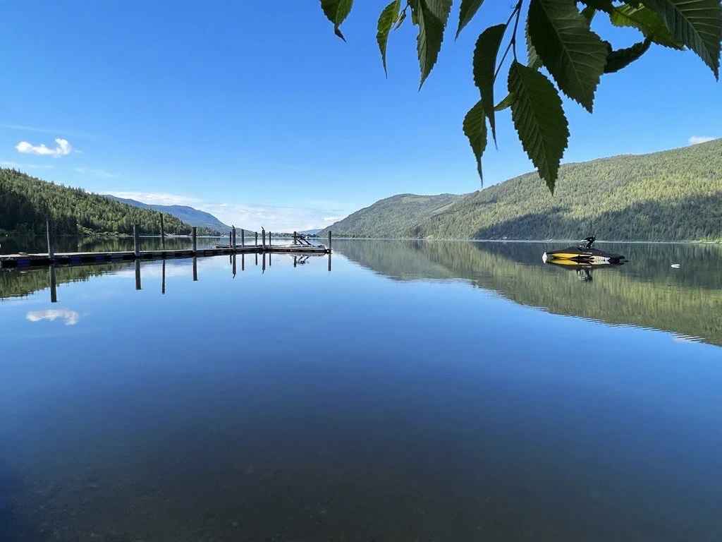A calm lake surrounded by green mountains under a clear blue sky, with a dock extending into the water and a boat on the right side.