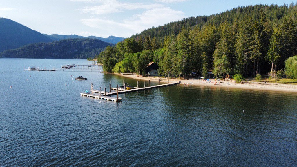 A lakeside scene with a wooden dock extending into calm water, surrounding by green trees and forested hills in the background.