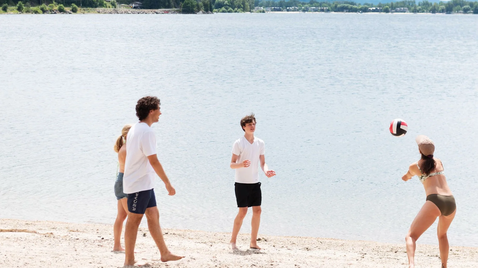 People playing volleyball on a sandy beach near the water, with a woman in a swimsuit hitting the ball while four others watch.