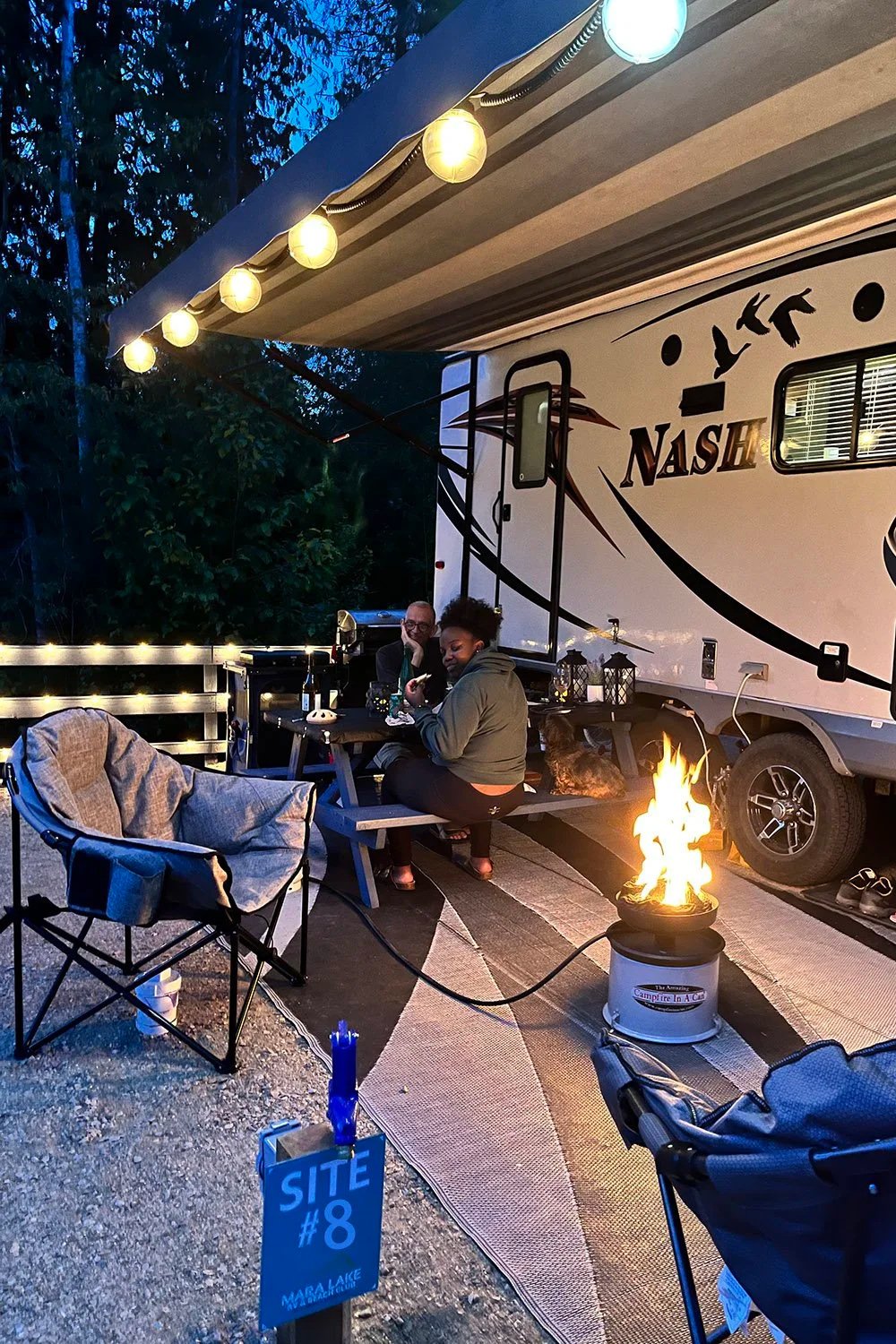 Campground with a large RV trailer labeled 'Nash' parked on a gravel site. There is a small fire pit lit with flames, and two people sitting at a picnic table near the trailer. One person is looking at their phone, and the other is sitting with their