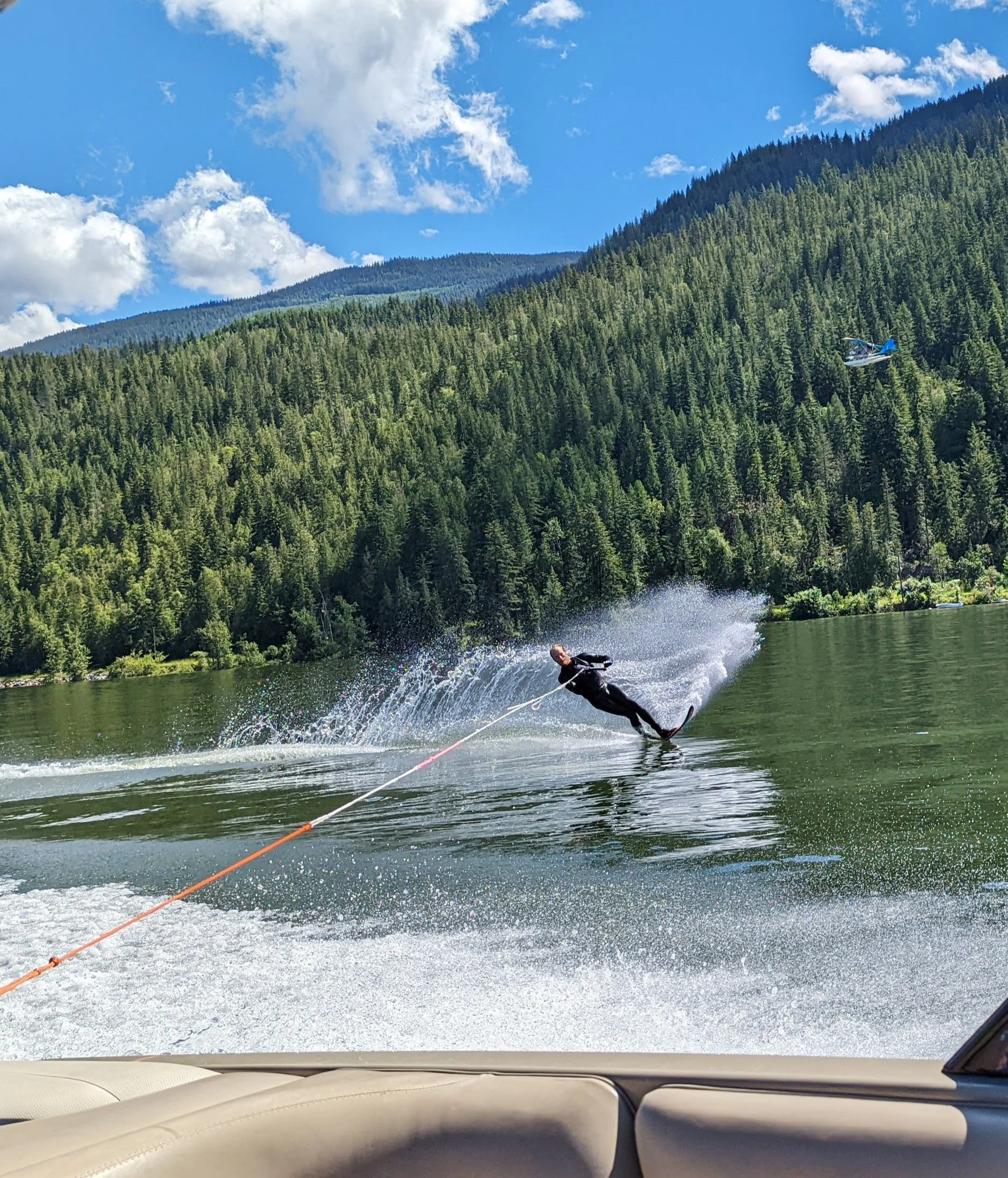 Person water skiing on a lake surrounded by lush green forest with pine trees, mountains in the background, and a partly cloudy blue sky.
