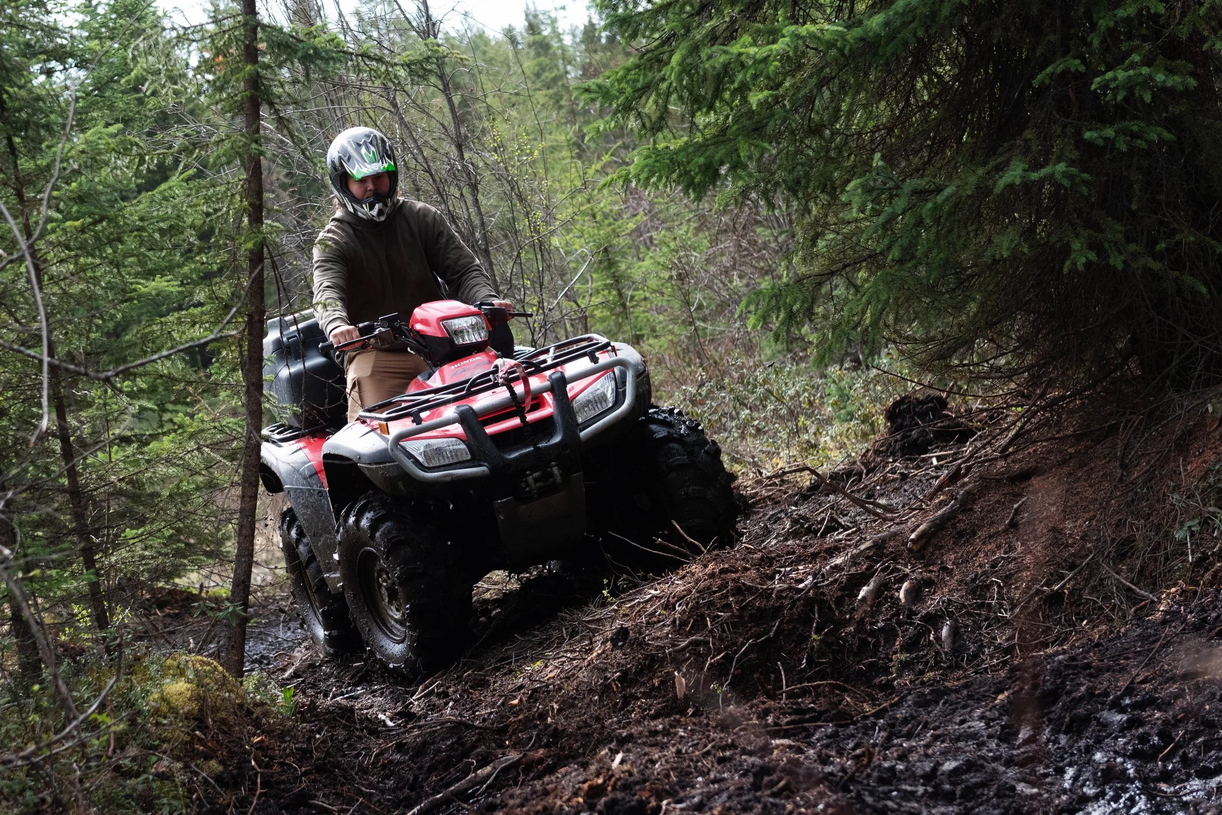 Person riding a red ATV through a muddy, wooded trail surrounded by green trees.