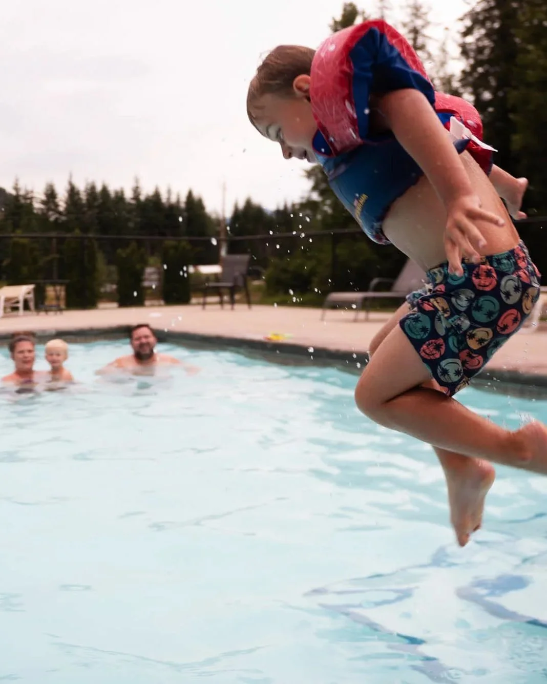 Child jumping into a swimming pool with family in the background, outdoors, on a cloudy day.