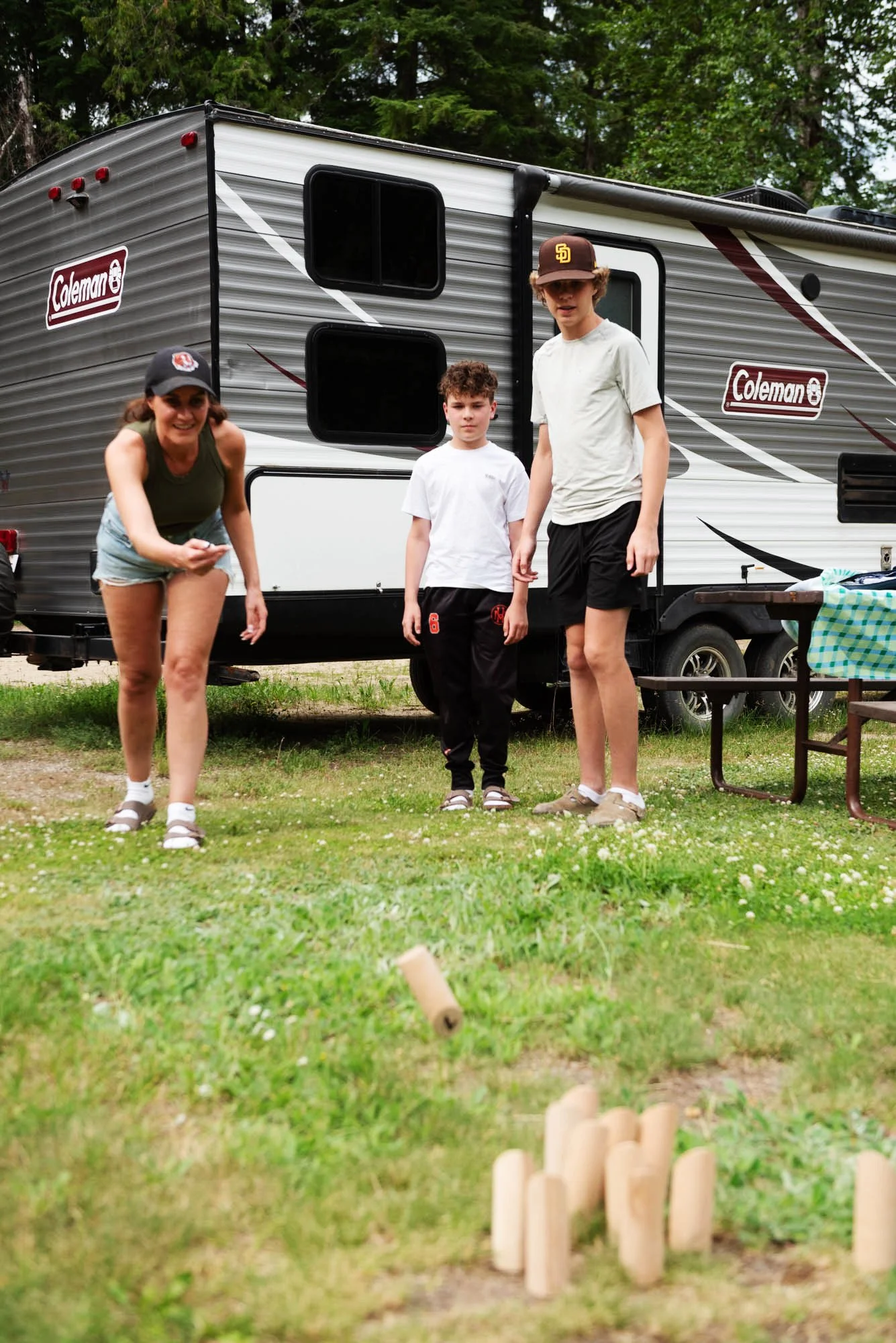 A woman and two boys standing outside near a travel trailer, with a small fenced area containing wooden sticks on the grass in foreground.