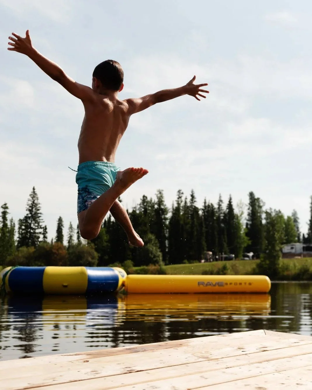 A boy mid-air jumping off a dock into a lake, with trees and a cloudy sky in the background.
