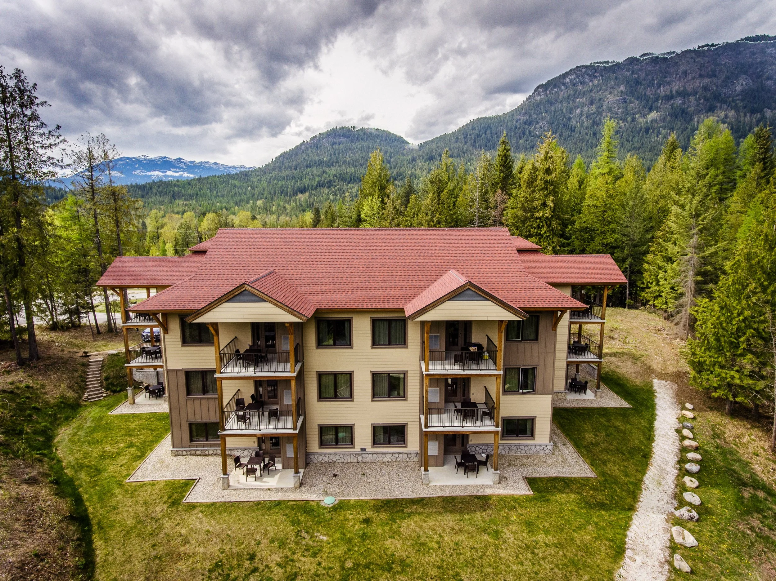 A multi-story residential building with balconies and outdoor furniture, set in a green, forested area with mountains and cloudy skies in the background.