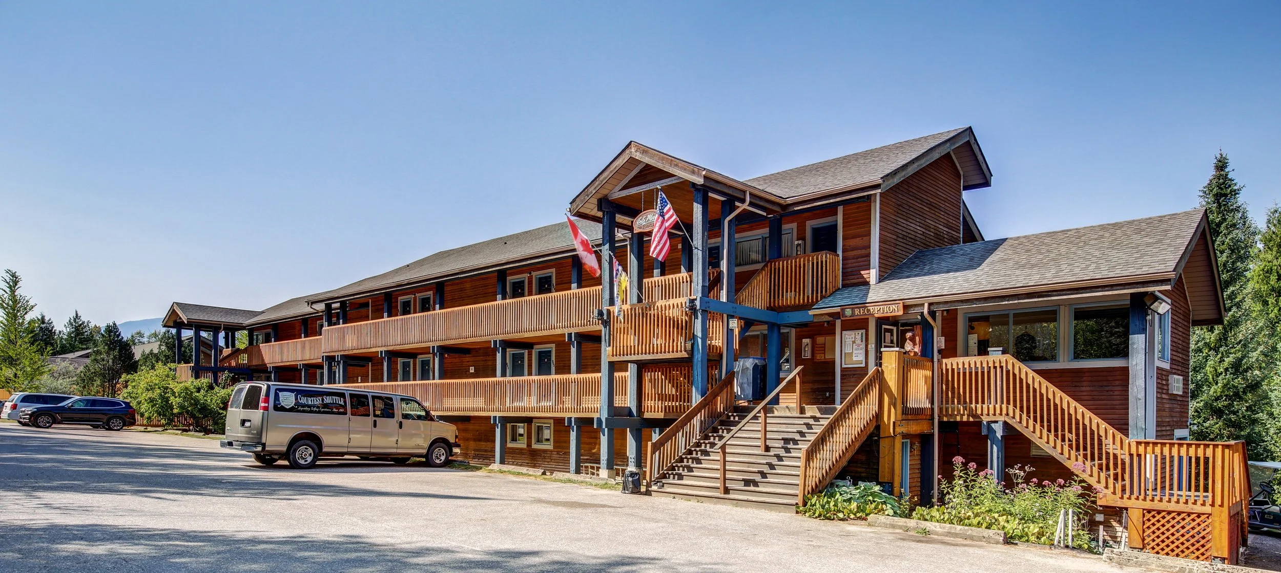 A multi-story wooden hotel with a staircase leading to the entrance, flags hanging on the porch, a parking lot with cars, and surrounded by trees and mountains in the background.
