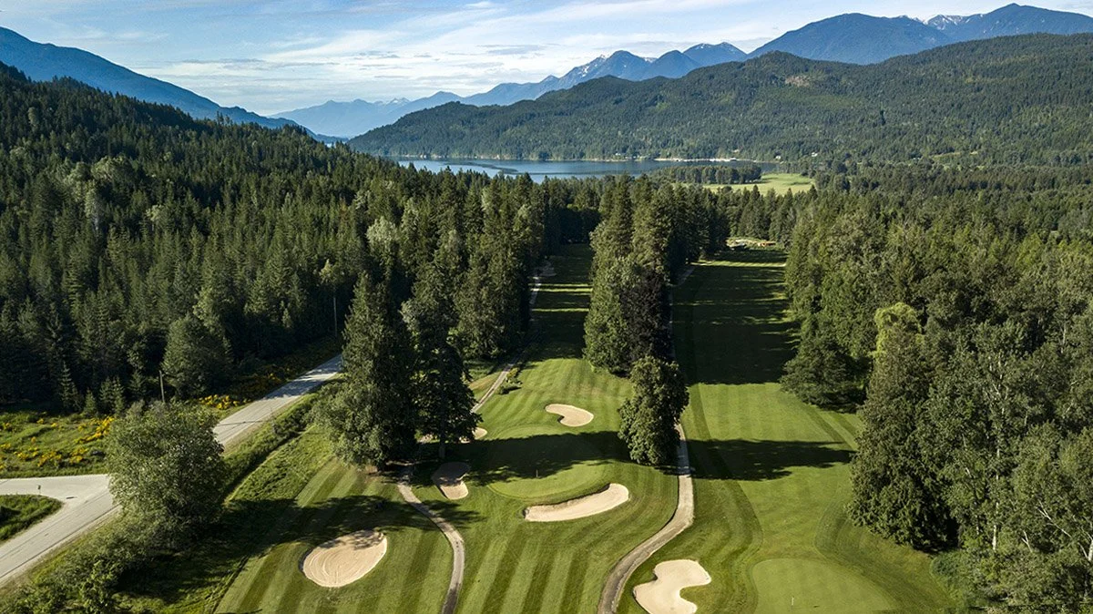 A scenic golf course surrounded by dense forest, with mountains in the background and a lake visible in the distance.