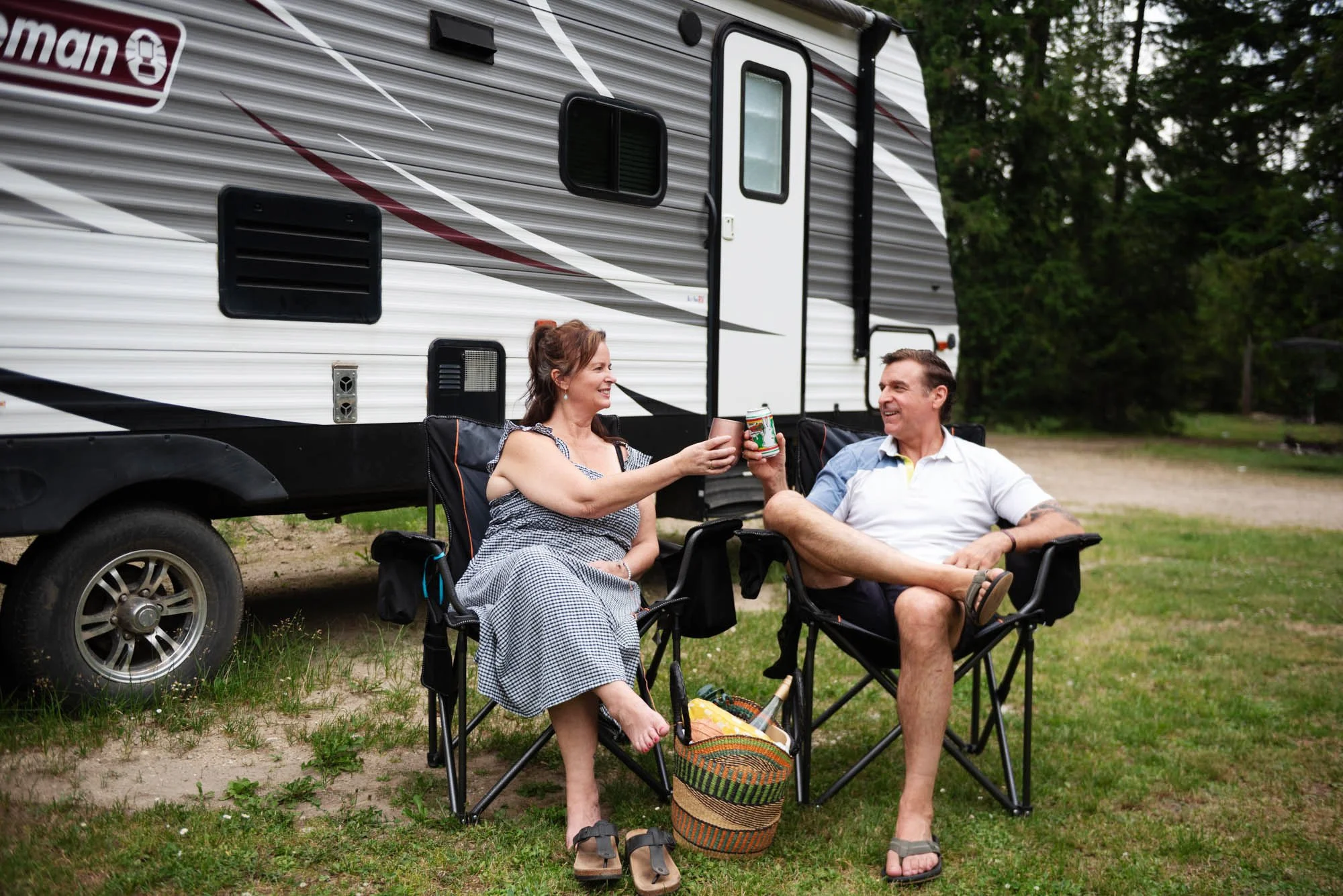 Two people, a woman and a man, sitting in camping chairs outdoors in front of a camper trailer. They are smiling and toasting with drinks, enjoying a casual moment in a wooded area.