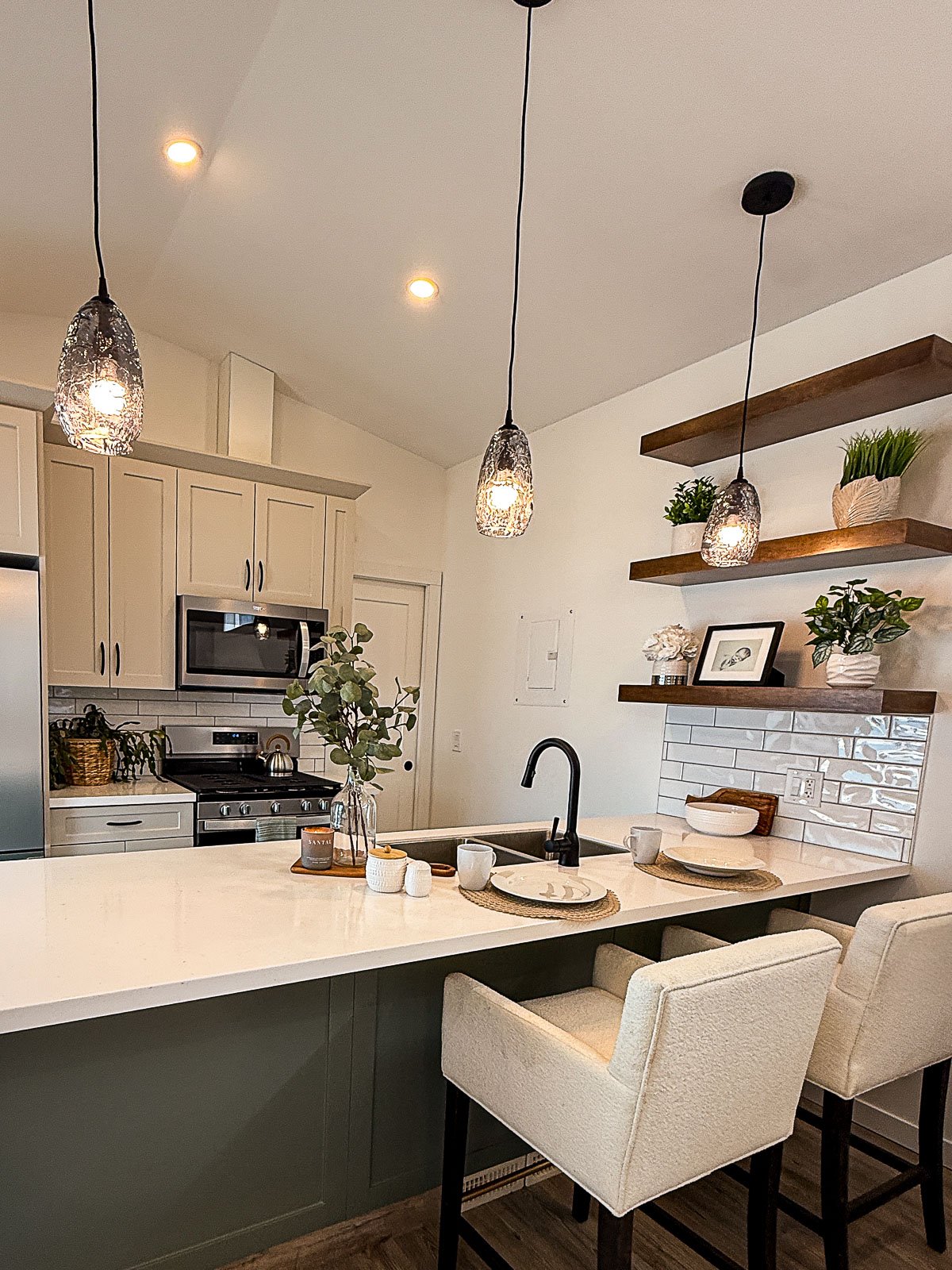 Modern kitchen with white cabinetry, black fixtures, open wooden shelves with plants and framed art, white island with seating, and pendant lighting.
