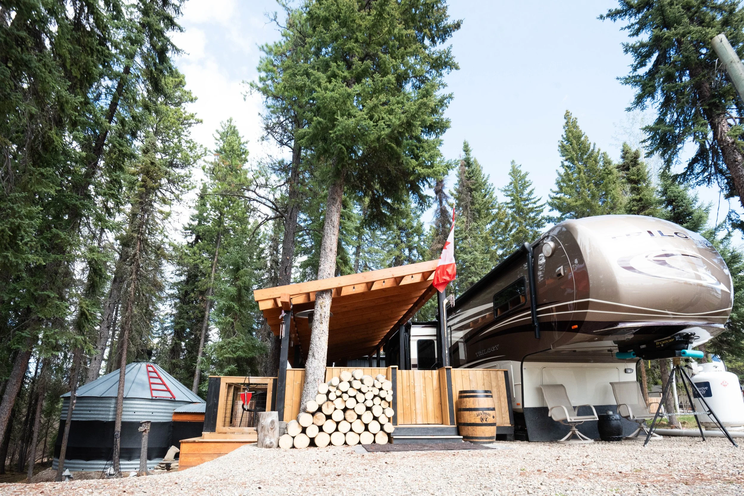 A forested outdoor scene with tall green pine trees, a small metal shed, a wooden deck, a large RV with a slide-out, a propane tank, a log pile, and a gravel ground.