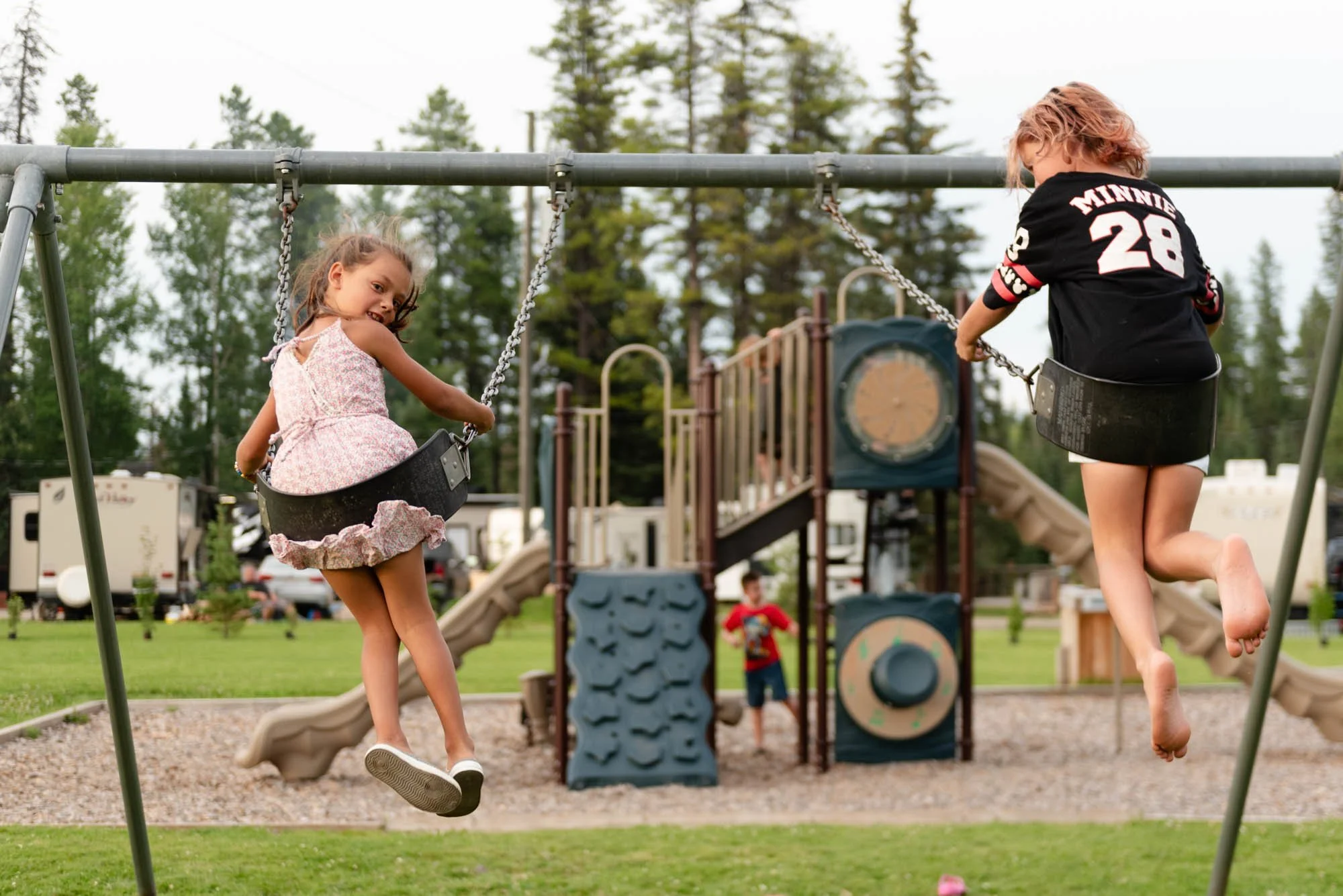 Two children swinging on swings at a playground, with slides and trees in the background.