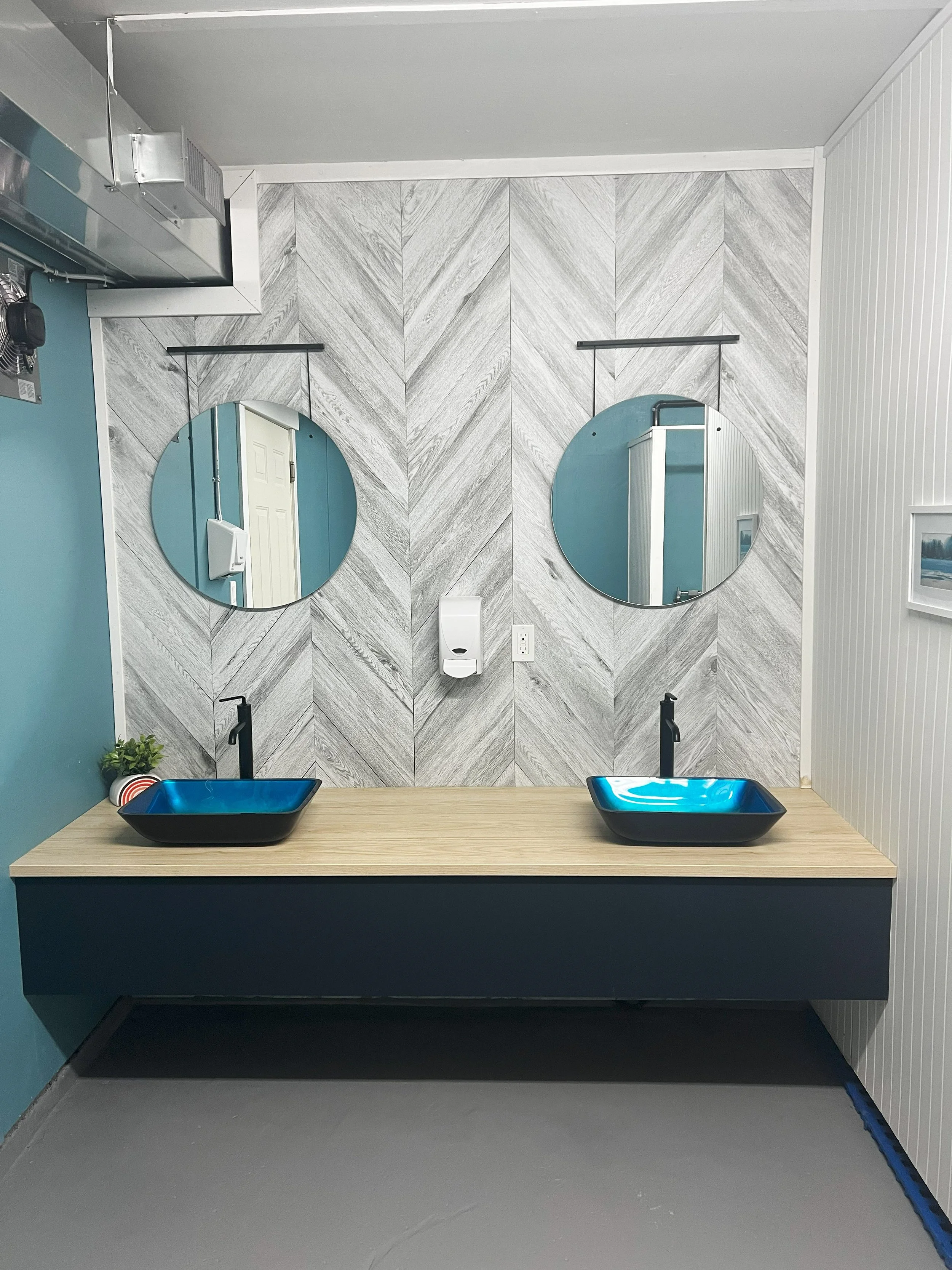 Public restroom with two mirrors, two black sinks with blue interiors, a hand sanitizer dispenser, and a small plant on the wooden countertop. The wall features a gray herringbone pattern.