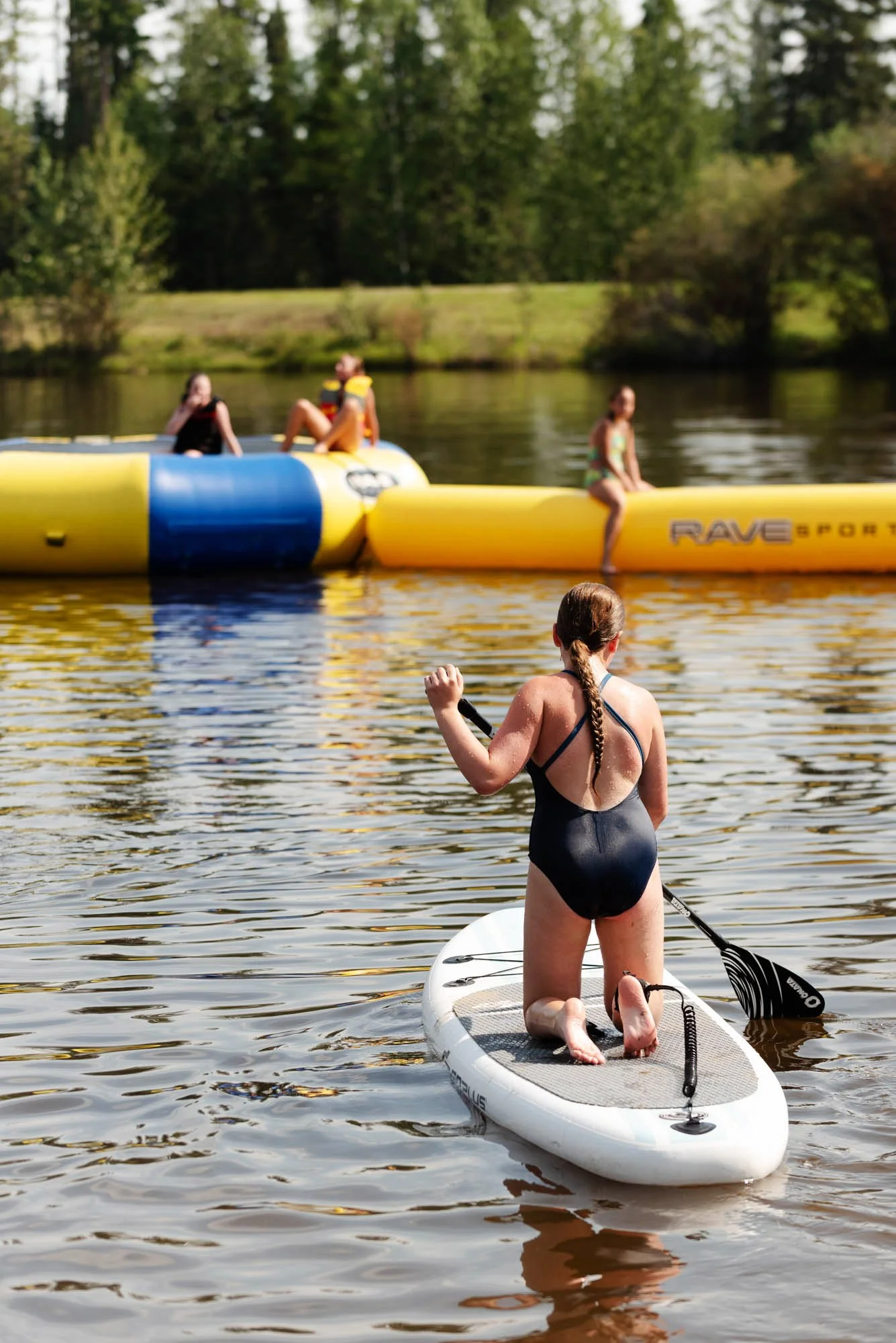A woman in a black swimsuit kneeling on a paddleboard, holding a paddle, on a calm lake, with a woman sitting on a yellow inflatable watercraft and another woman sitting on the yellow inflatable watercraft in the background, surrounded by green trees