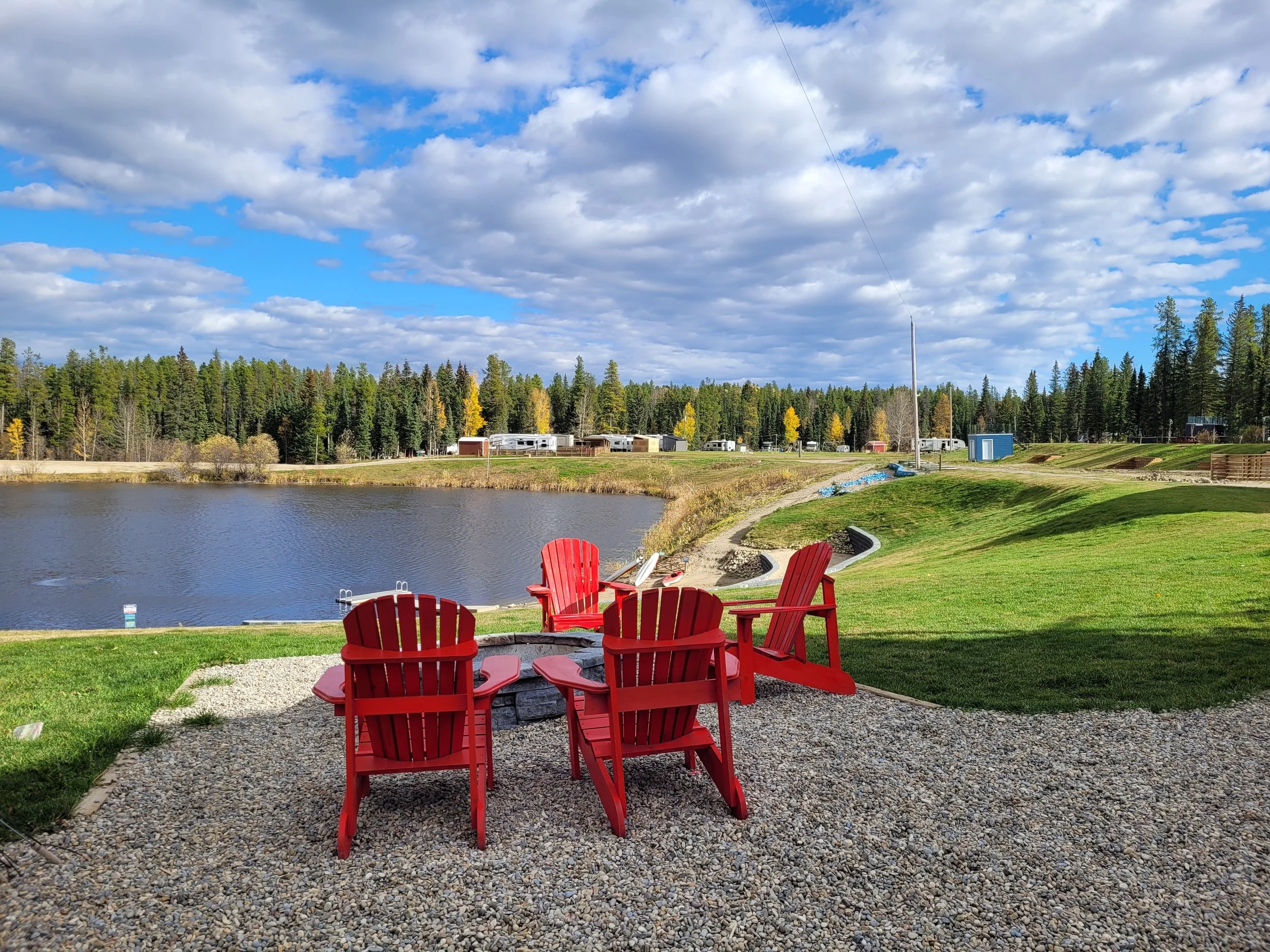 Red outdoor chairs arranged around a fire pit near a lake with trees and a partly cloudy sky in the background.