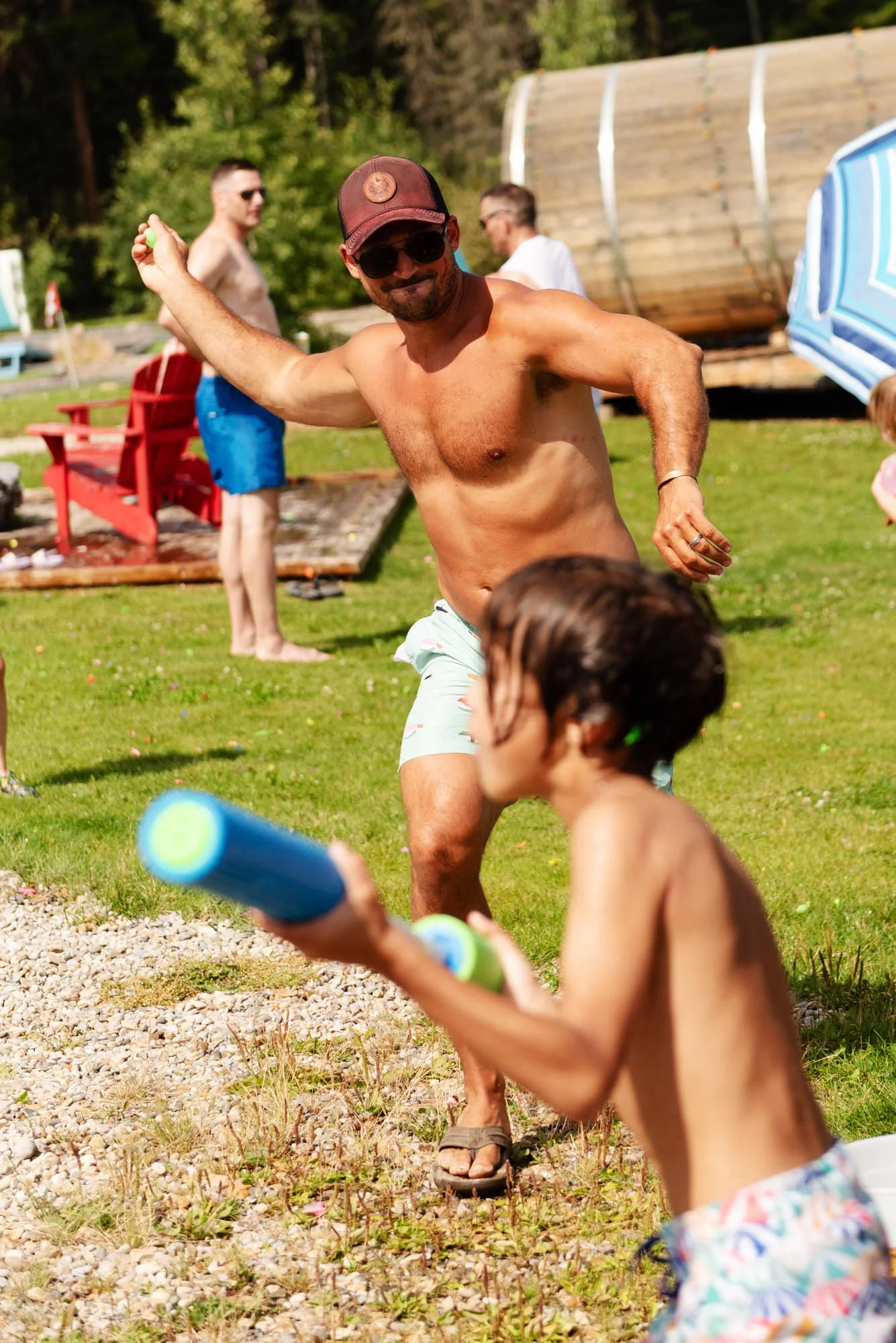 A shirtless man with sunglasses and a cap is smiling and dancing outdoors near children holding foam water toy sticks, with other people and a wooden structure in the background.