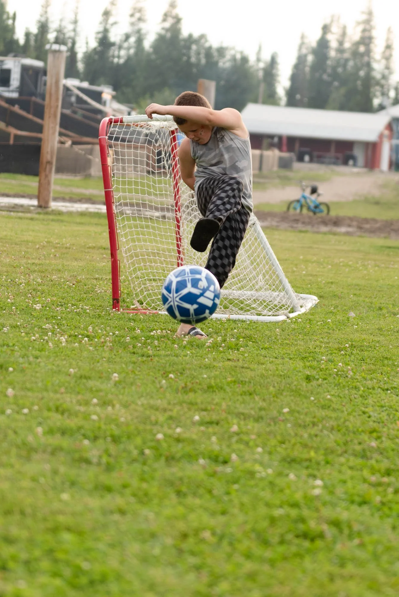 A young boy playing soccer outdoors during daytime, kicking a ball towards a small goal.