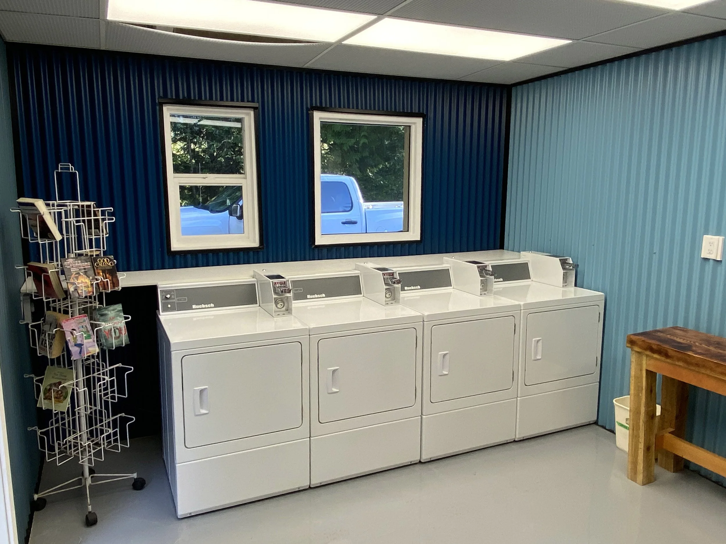 Interior of a laundromat with four white coin-operated washing machines lined up against a dark blue wall. To the left, a white wire rack displays magazines and postcards. On the right, a small wooden table is visible. Two windows are on the wall, sh