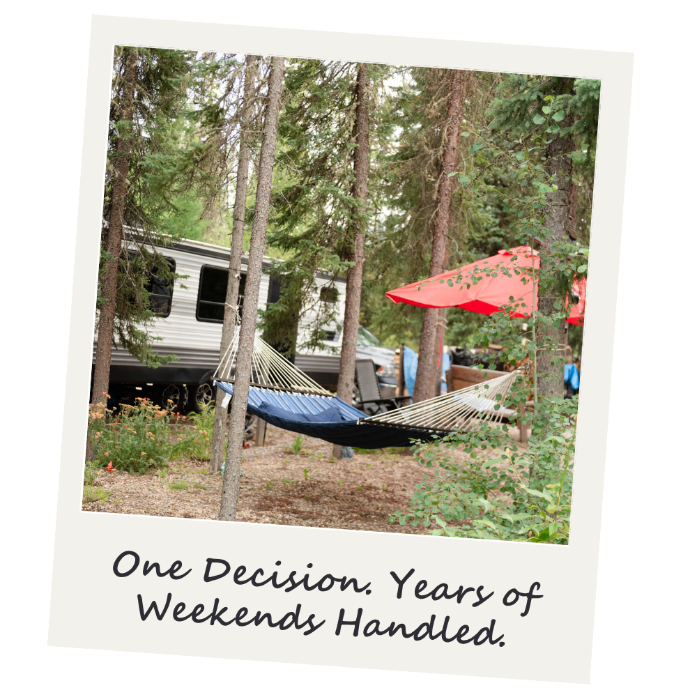A hammock tied between two trees in a wooded area with a camper, orange umbrella, and camping chairs in the background.
