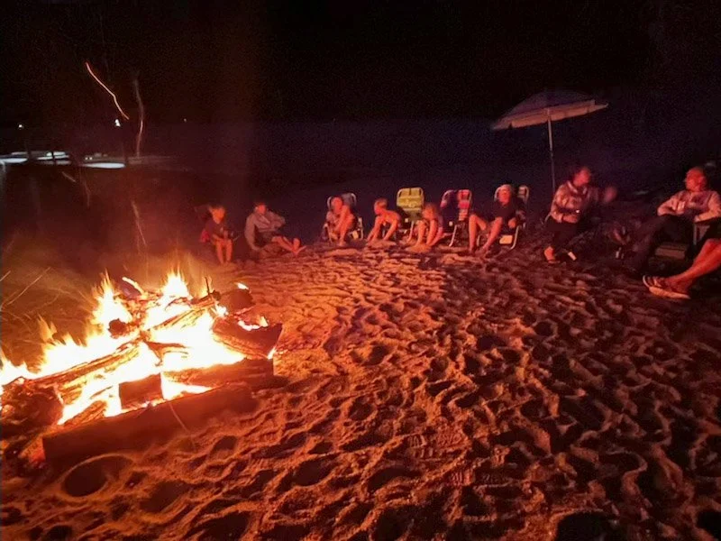 People sitting on chairs around a campfire on the sandy beach at night.