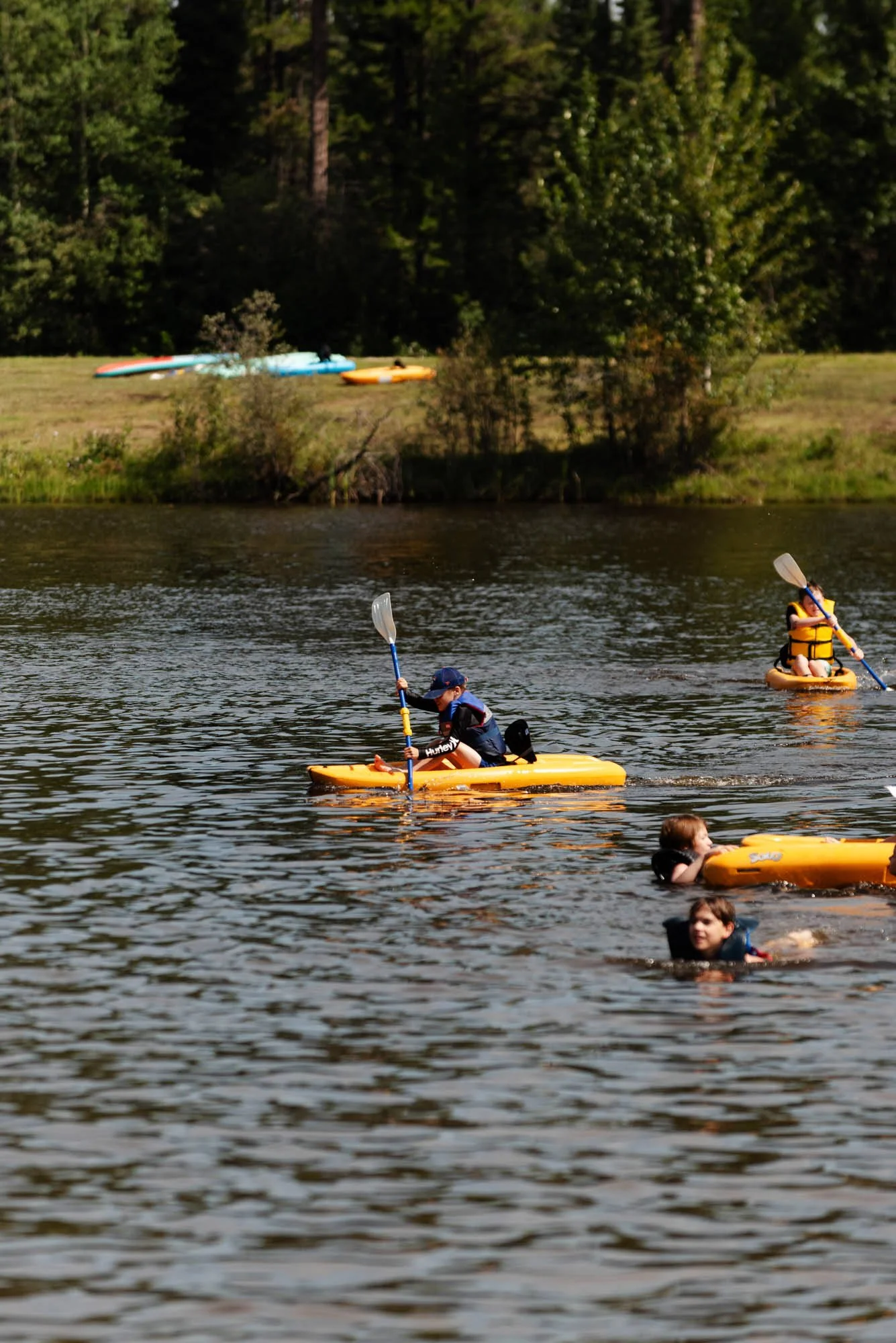 Children and adults kayaking and swimming in a lake with a grassy shoreline and trees in the background.