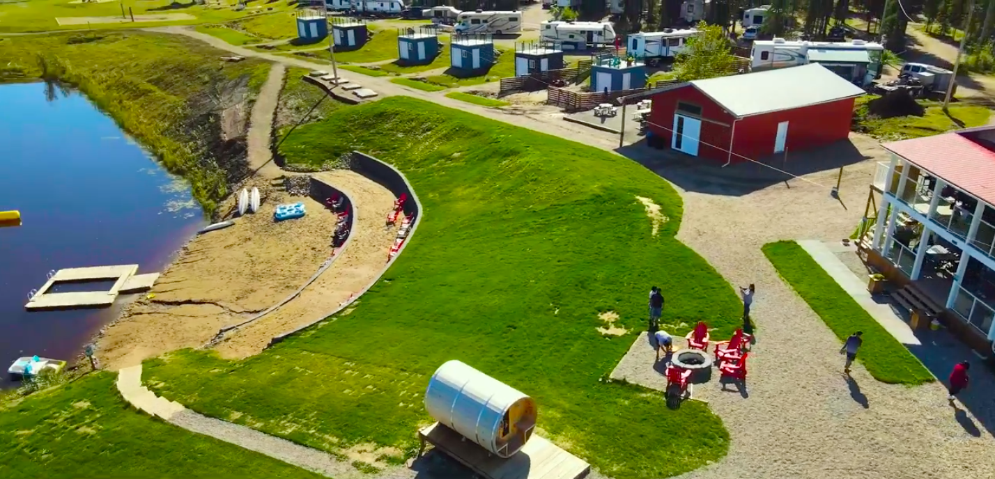 A lakeside campground with sand volleyball court, small boats docked on the shore, RVs, a red building, a fire pit with chairs, and people walking and socializing on the grassy and gravel areas.