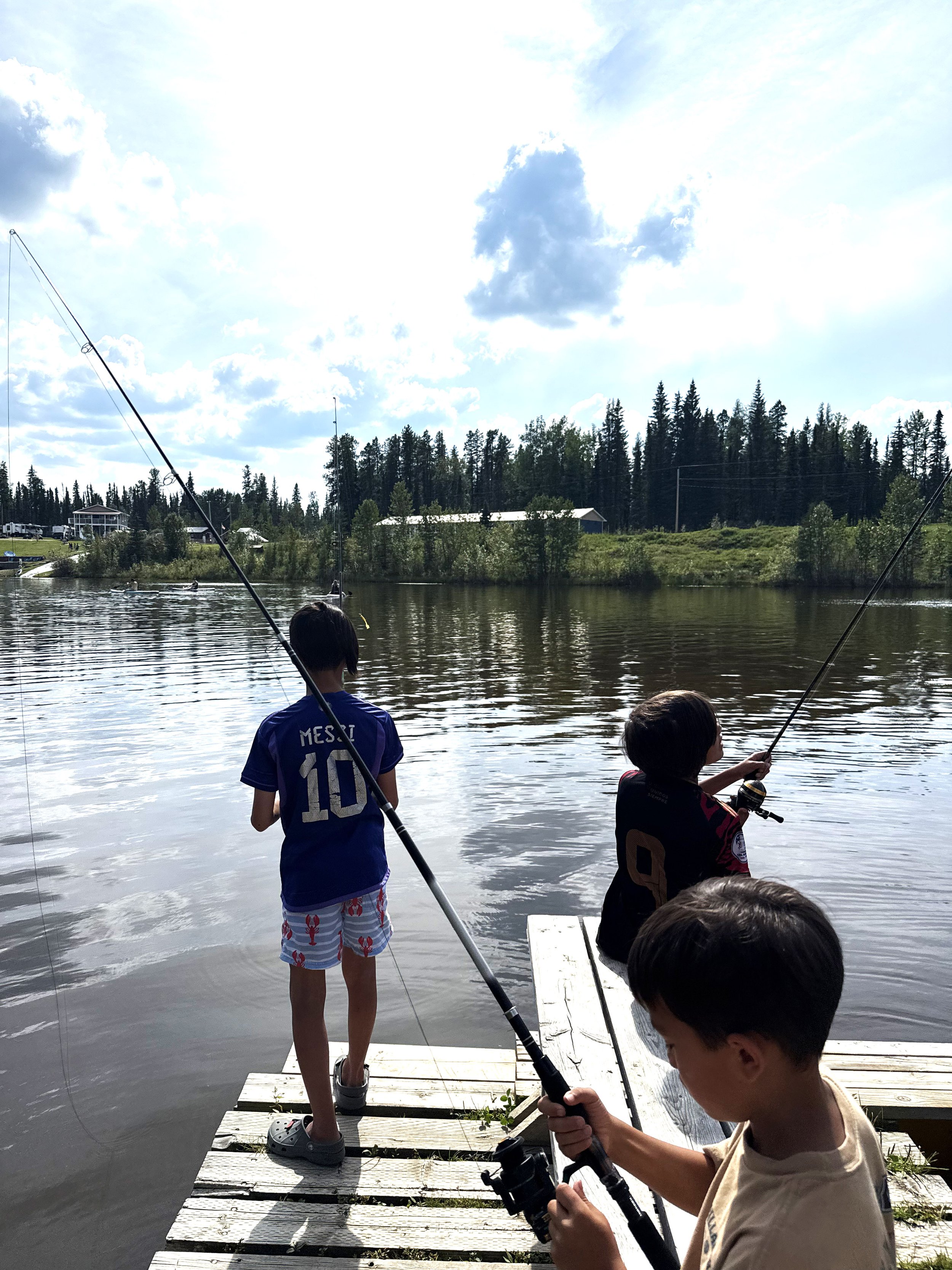 Children fishing off a dock by a lake, with trees and a building in the background under a partly cloudy sky.