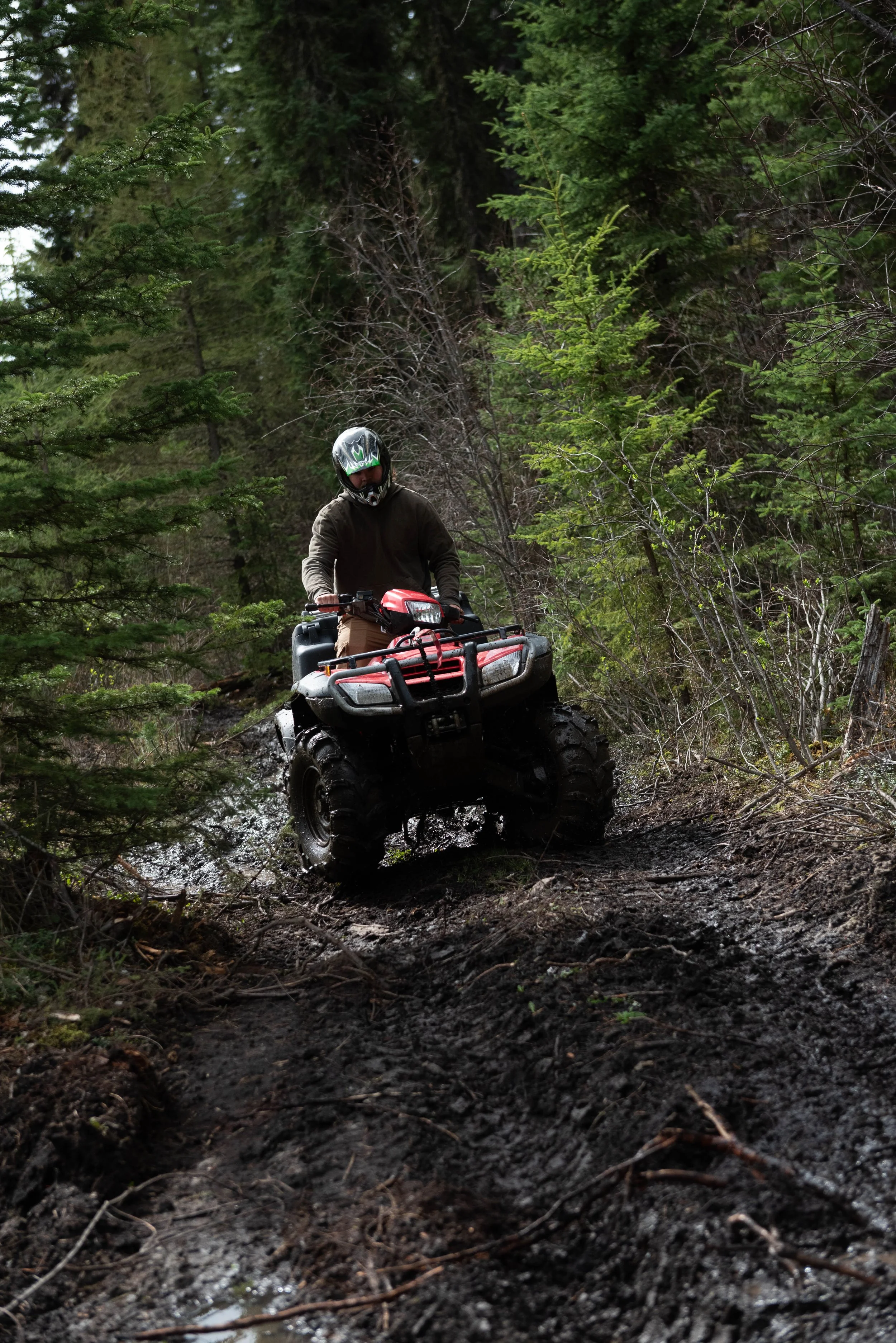 A person in a black helmet and brown jacket riding a red all-terrain vehicle (ATV) through a muddy forest trail surrounded by greenery.