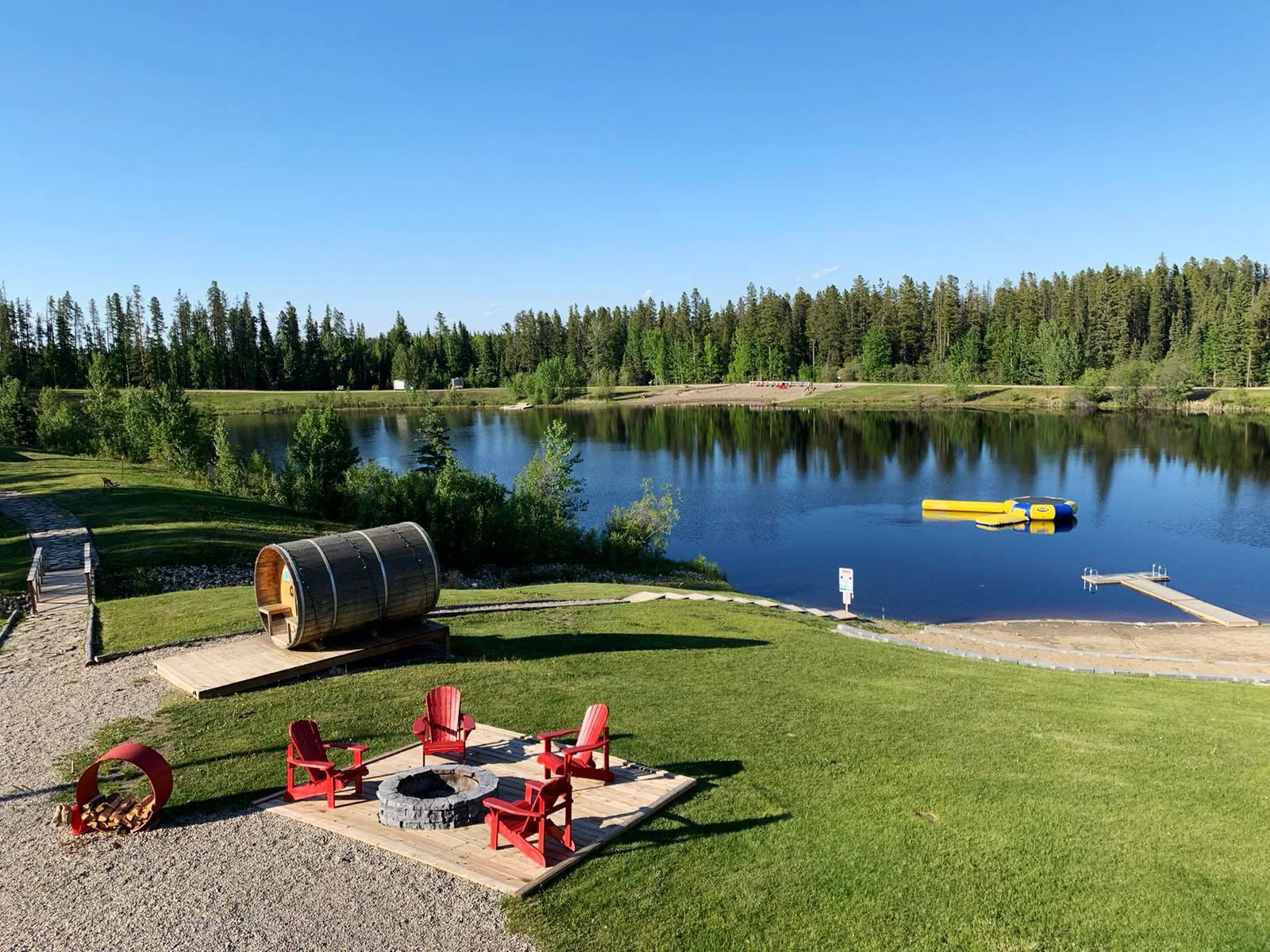View of a calm lake surrounded by green trees, with a fallen floatation device and a dock on the shore, and red chairs and a fire pit in the foreground.