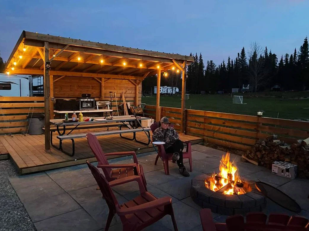 An outdoor patio area with a fire pit, red chairs, and a wooden covered deck with string lights at dusk. There is a person sitting with a phone near the fire, and a stack of firewood on the side.
