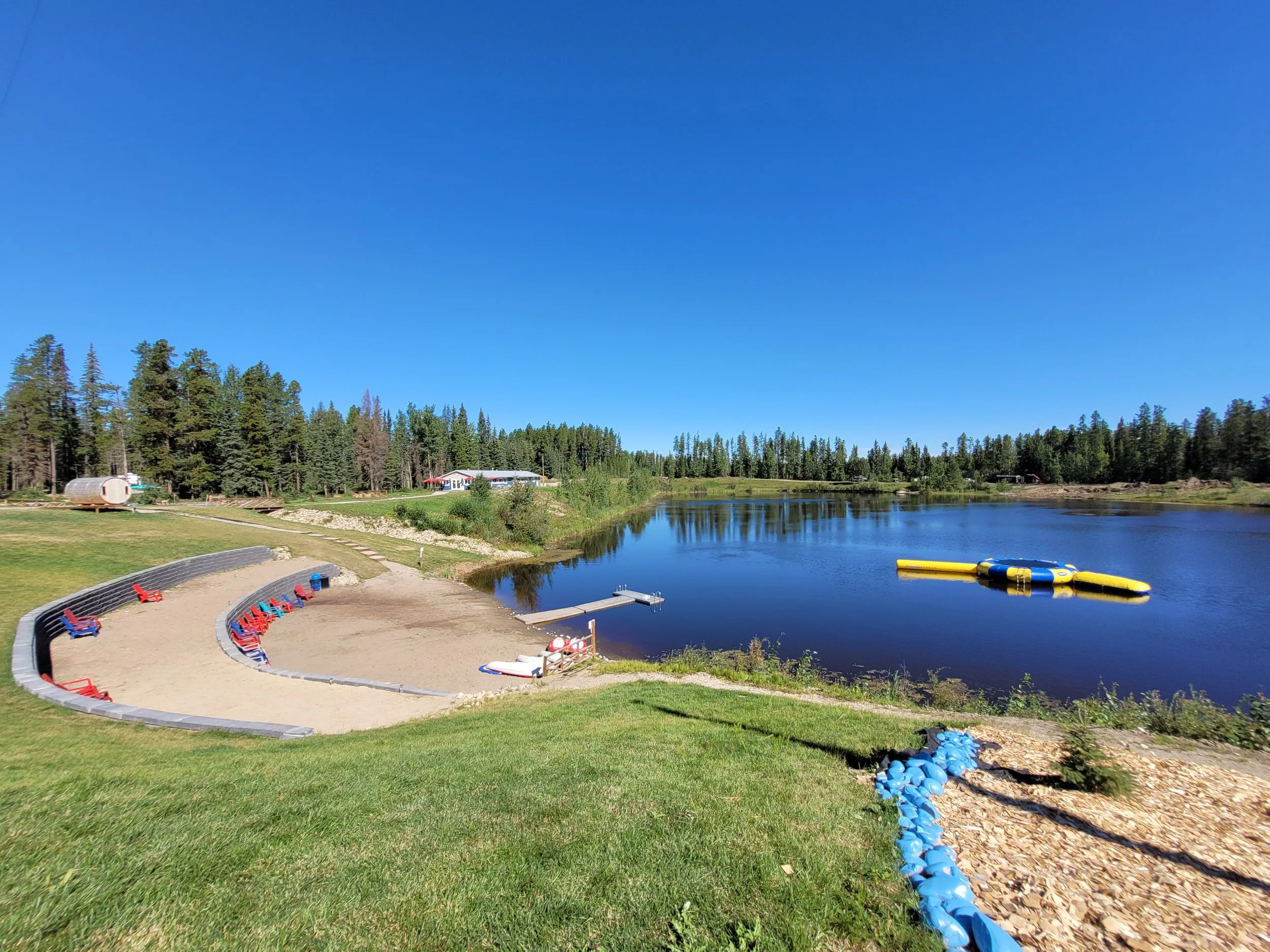 A lakeside scene with a clear blue sky, trees, a building, and inflatable water toys on the lake, with a sloped grassy area and blue plastic balls.